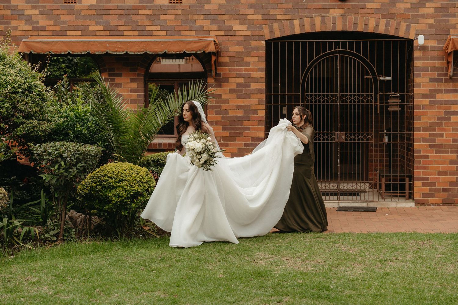 Bride in white gown with bridesmaid in olive dress holding train outside brick building with lush garden.