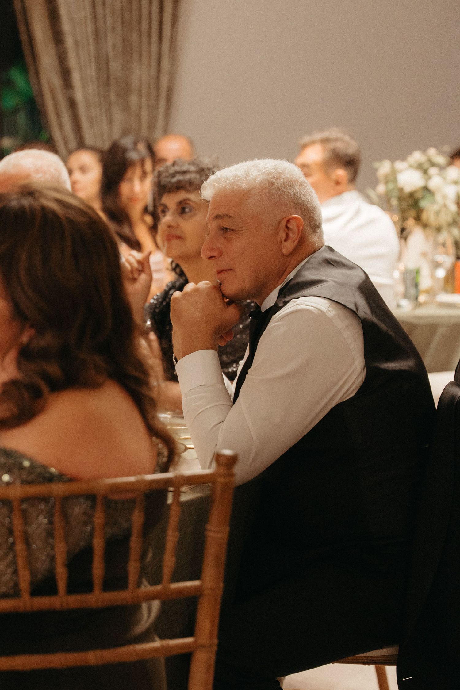 Elderly man in vest and bow tie sits pensively at a wedding reception, surrounded by guests in elegant attire.