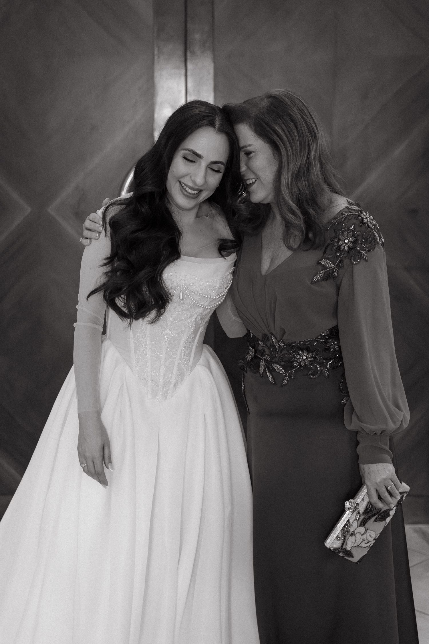 Black and white photo of a smiling bride in a white gown embracing a woman in a beaded dress at a wedding.