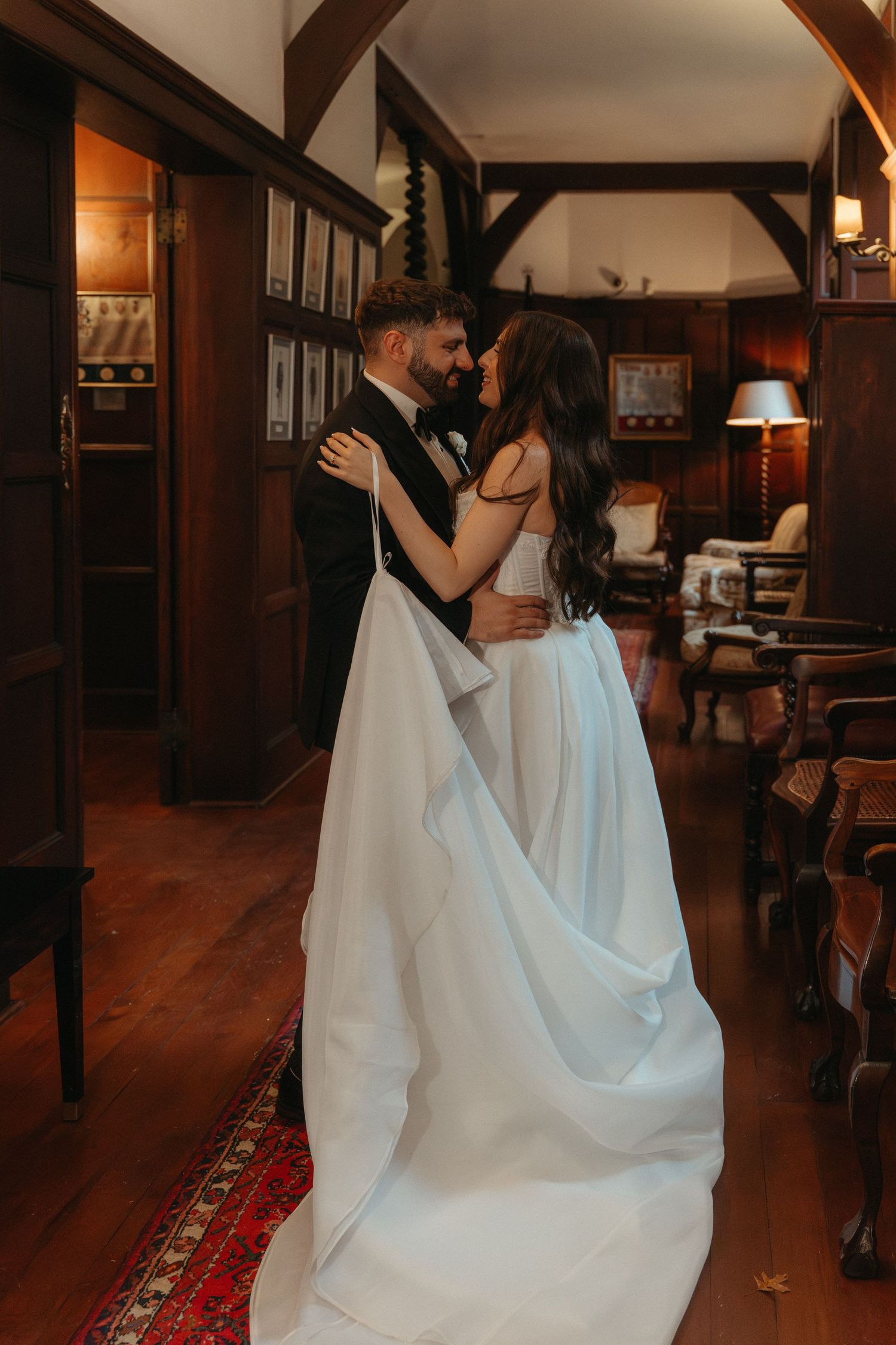 Bride and groom share a romantic embrace in an elegant wood-paneled room, bride wearing a flowing white gown.