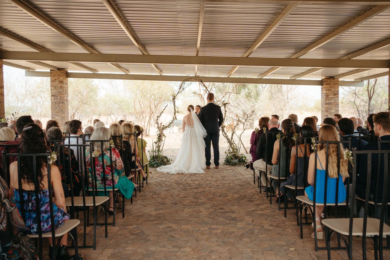 Wedding ceremony taking place under a rustic covered outdoor venue with guests seated on dark chairs.