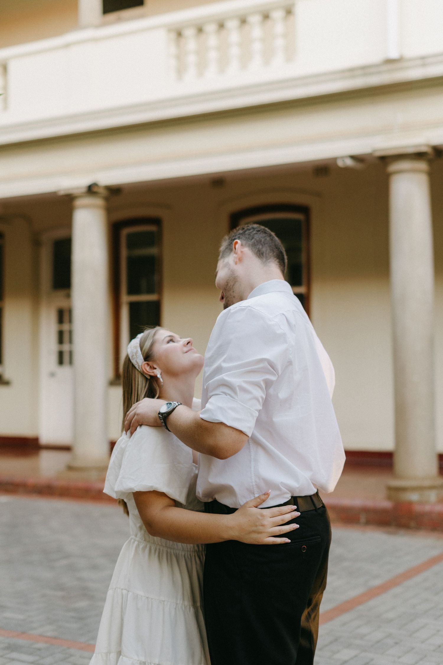 A tender embrace between a couple wearing white and black outfits in front of a colonial-style building.