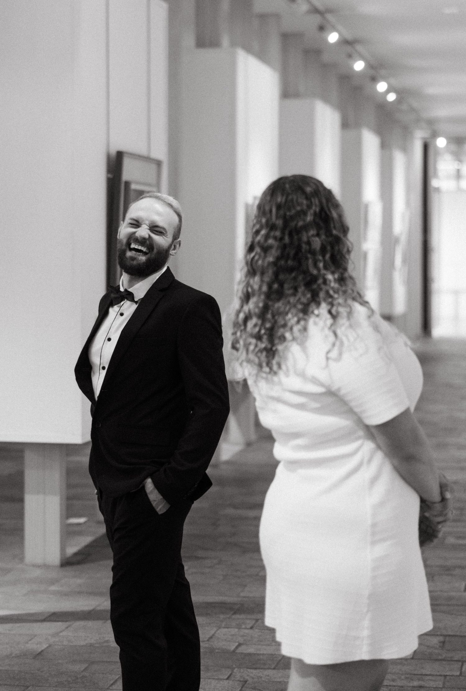 Joyful moment captured in black and white as couple laughs in elegant gallery hallway.