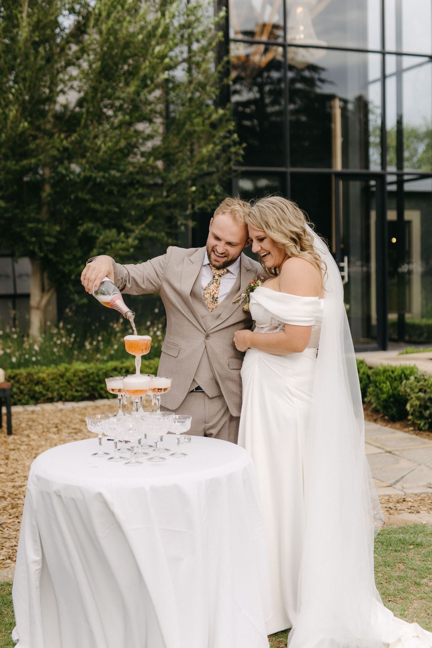 A couple pours champagne into a tower of glasses during an elegant outdoor wedding celebration.