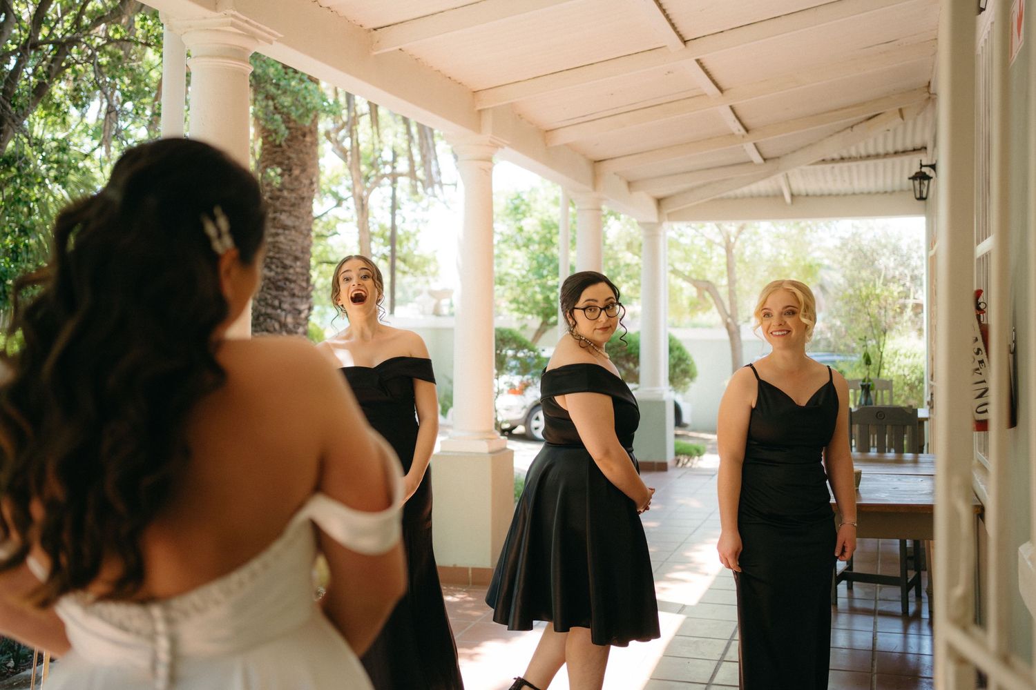 A group gathers in formal attire on a covered porch with natural light streaming through the columns.