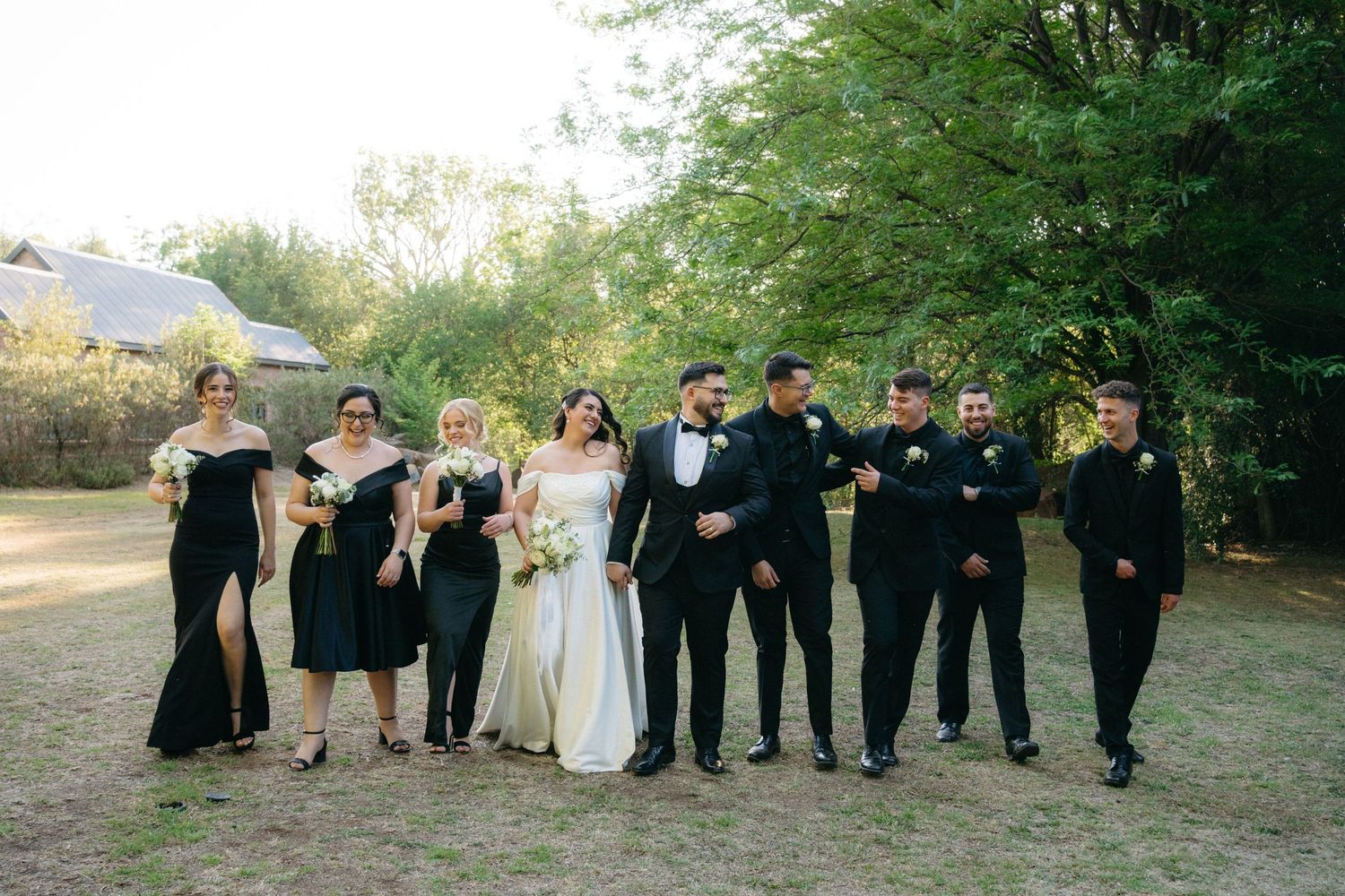 Wedding party in formal black and ivory attire poses together outdoors on a sunny day with trees in background.