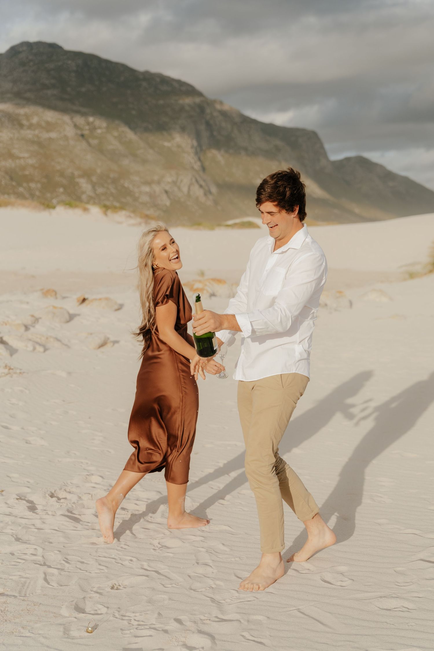 Couple dancing barefoot at Betty's Bay at sunset, woman in brown satin dress holding champagne.