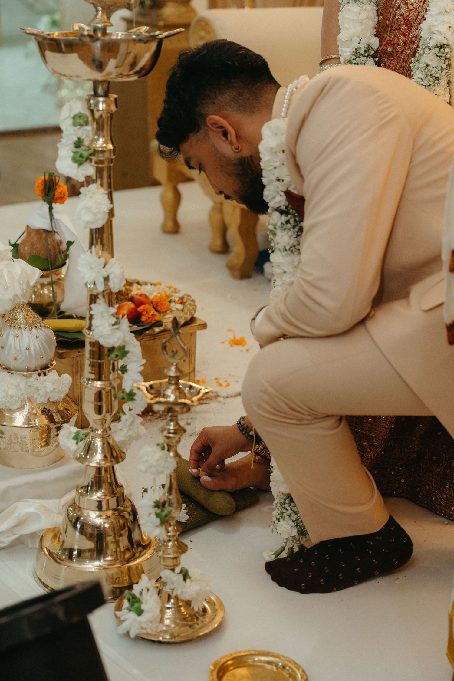 A person in a cream-colored suit kneels at an elegantly decorated table with silver candelabras and floral arrangements.