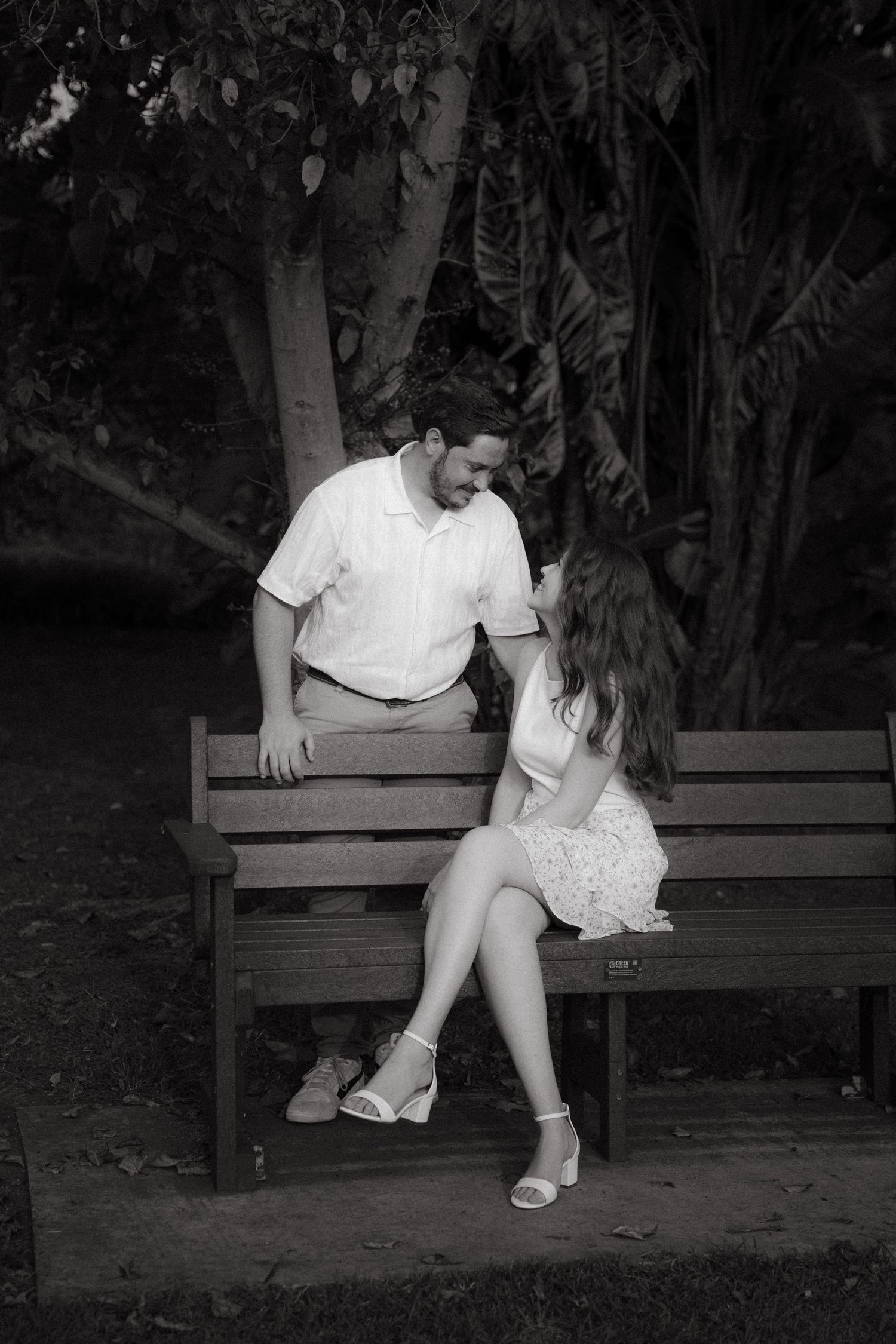 Black and white photo of a couple sitting on a park bench, sharing an intimate conversation surrounded by trees.