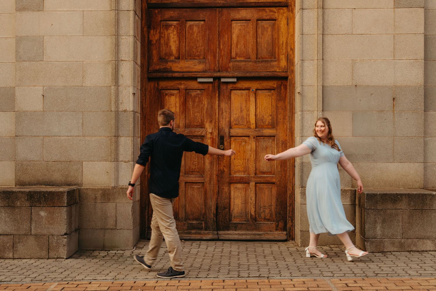 Two people dance playfully in front of a large wooden door on a stone building exterior.