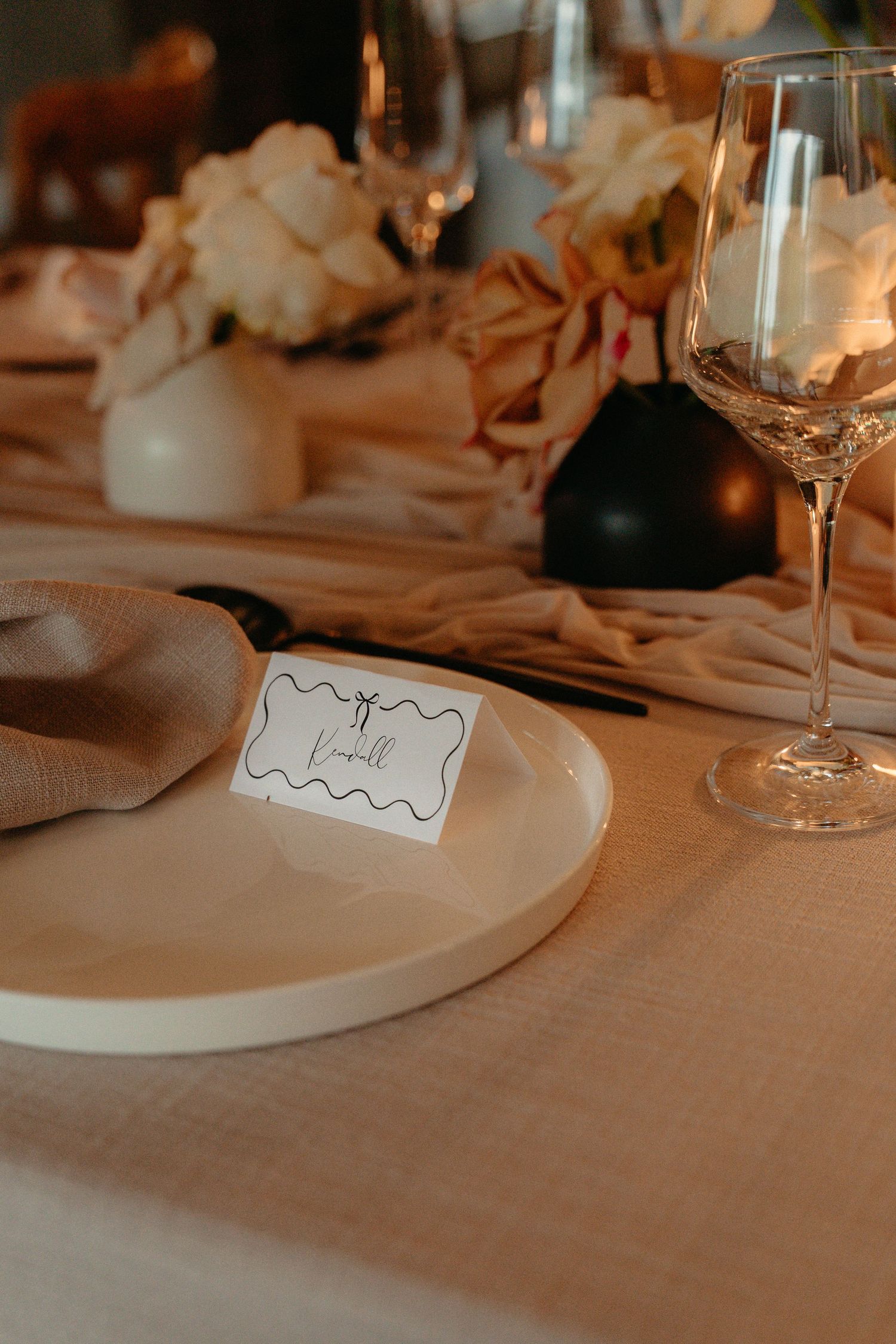 Elegant place setting at wedding reception with name card, white plate, and wine glass on beige tablecloth.
