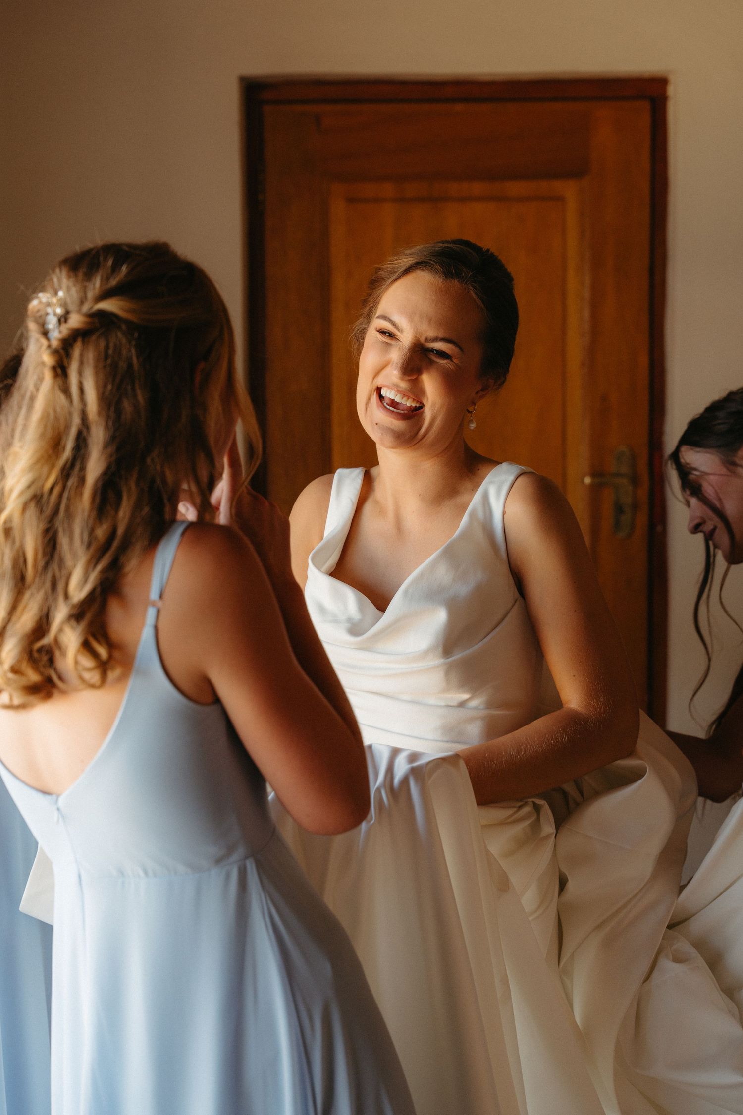 A group of bridesmaids in light blue and white dresses laugh together while getting ready for a wedding.