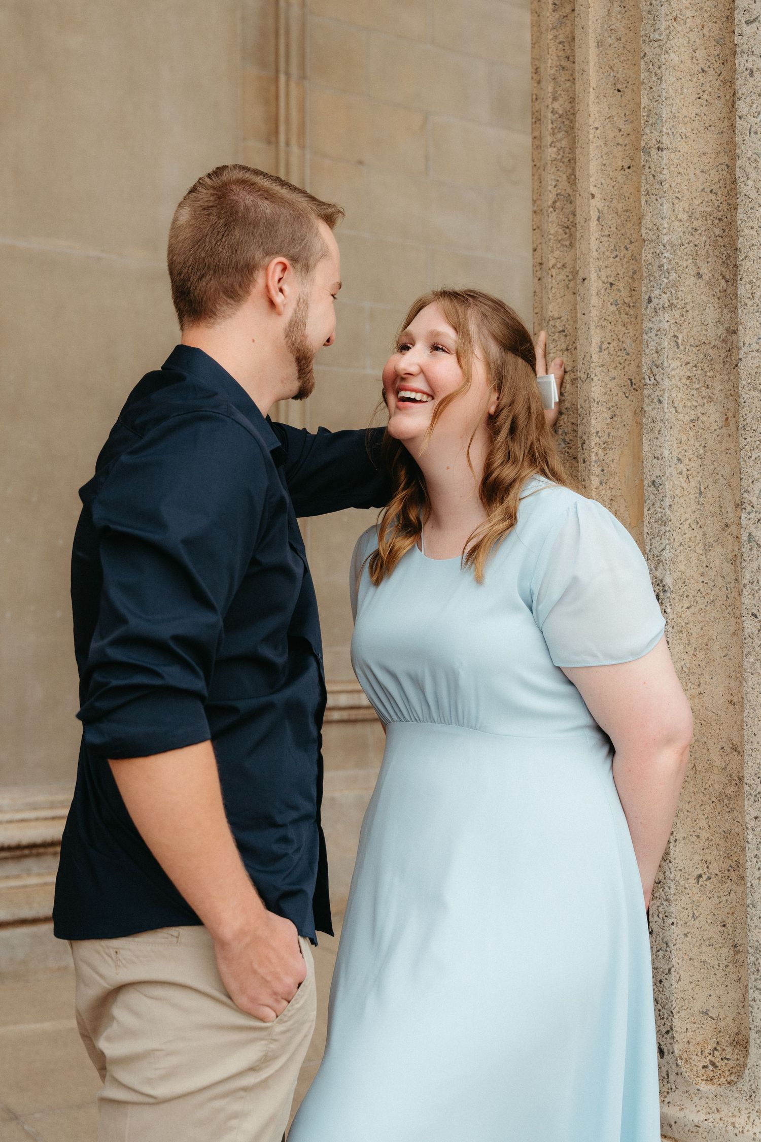 A couple shares a joyful moment together against a stone column backdrop, with her wearing a light blue dress.