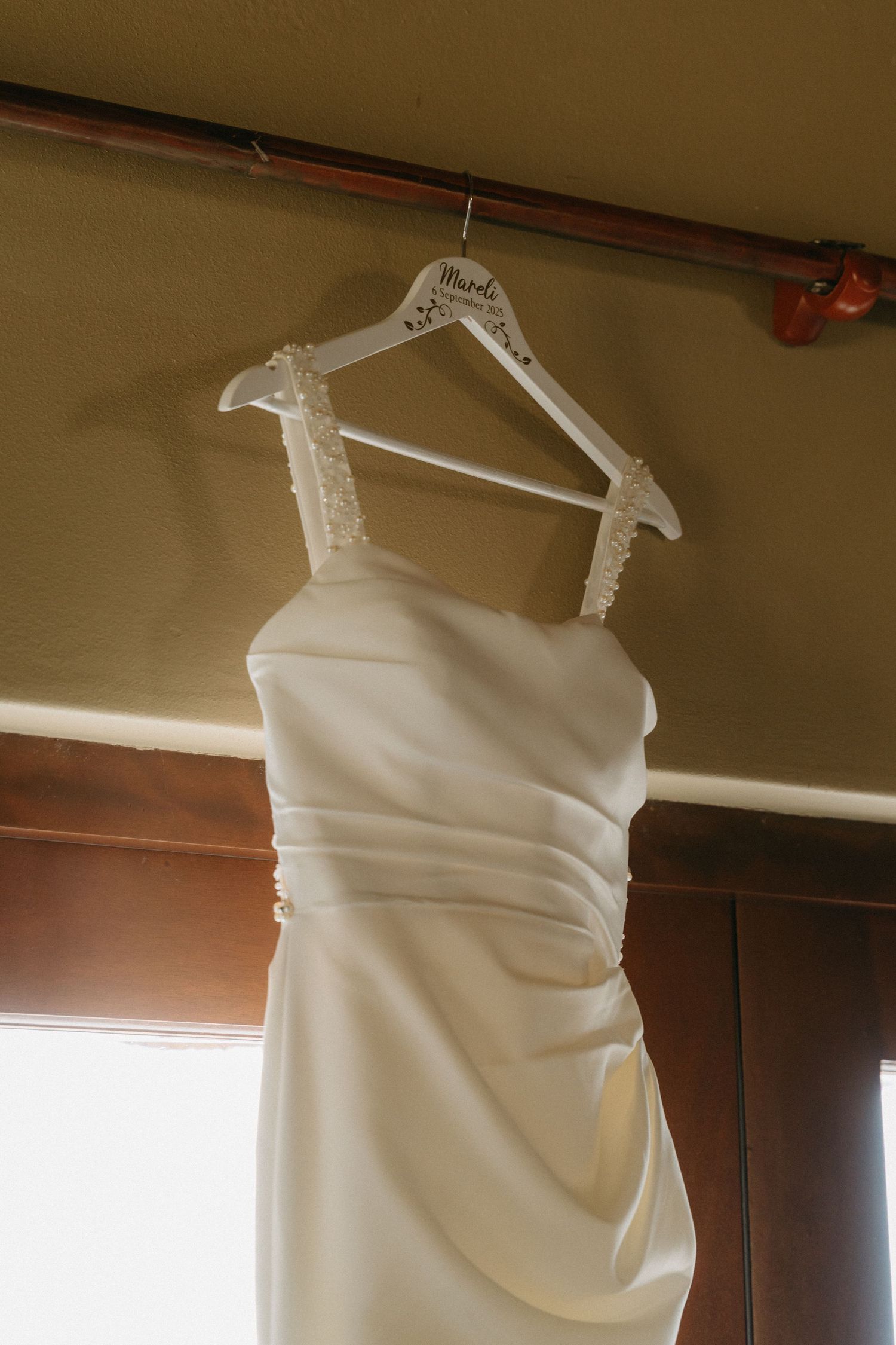 An elegant white wedding dress hanging on a custom hanger against a warm-toned wall.