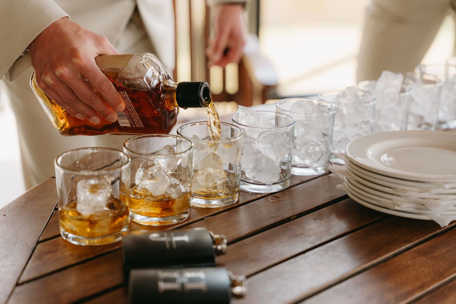 Hands pouring whiskey into glasses with ice at a wedding reception bar.
