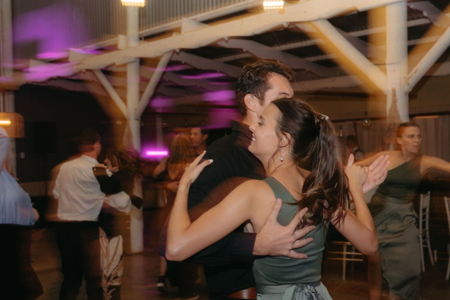 Guests dancing together at a dimly lit wedding reception with purple accent lighting.