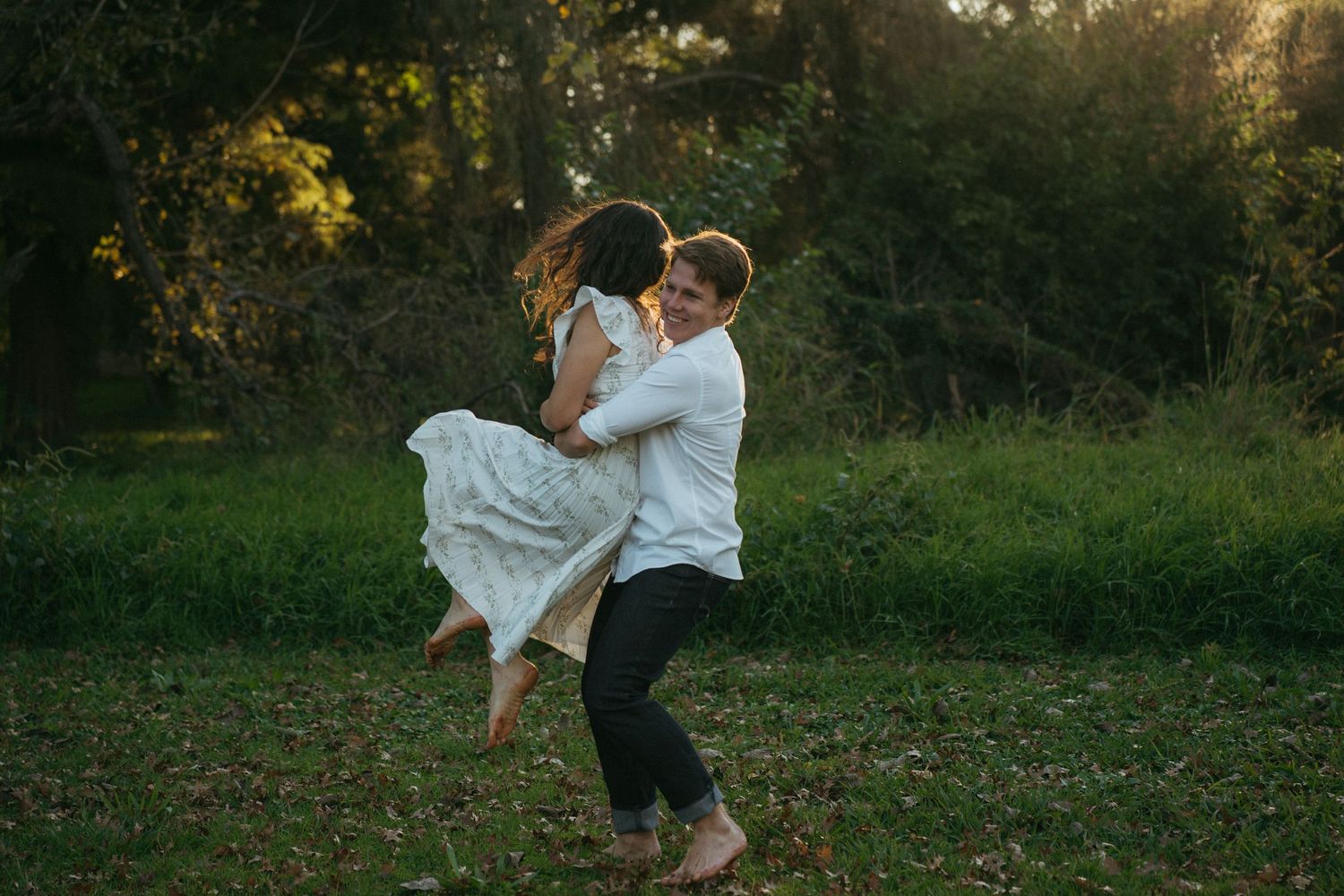 A couple dances together in a sunny meadow at golden hour, with the person in white being lifted and spun around.