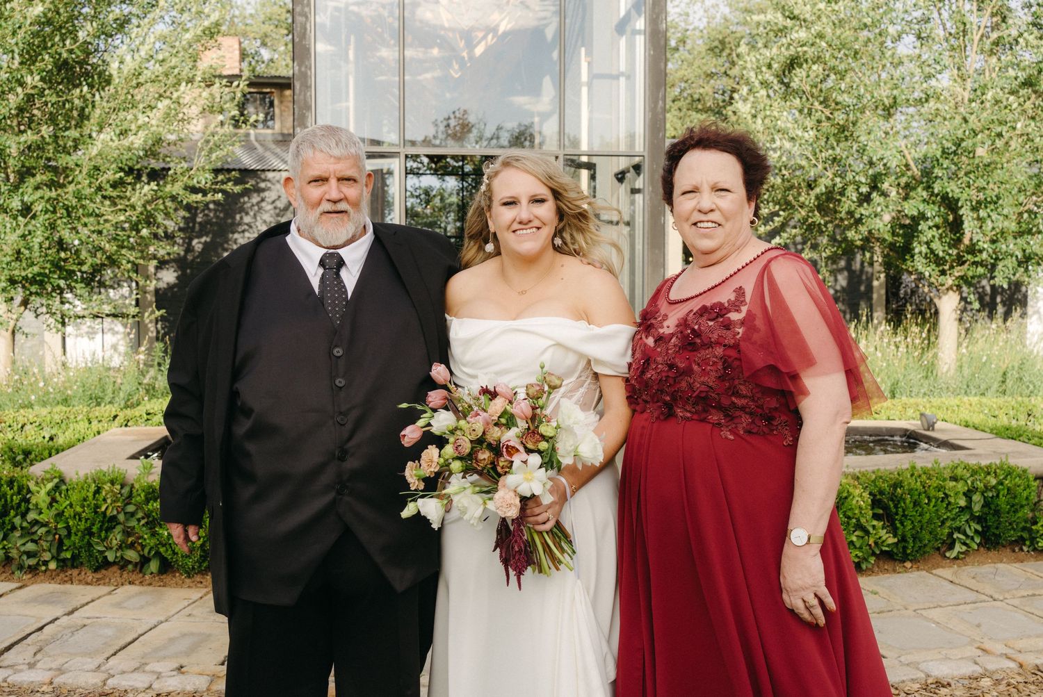 Wedding photo of three family members in formal attire standing in front of lush garden greenery on sunny day.