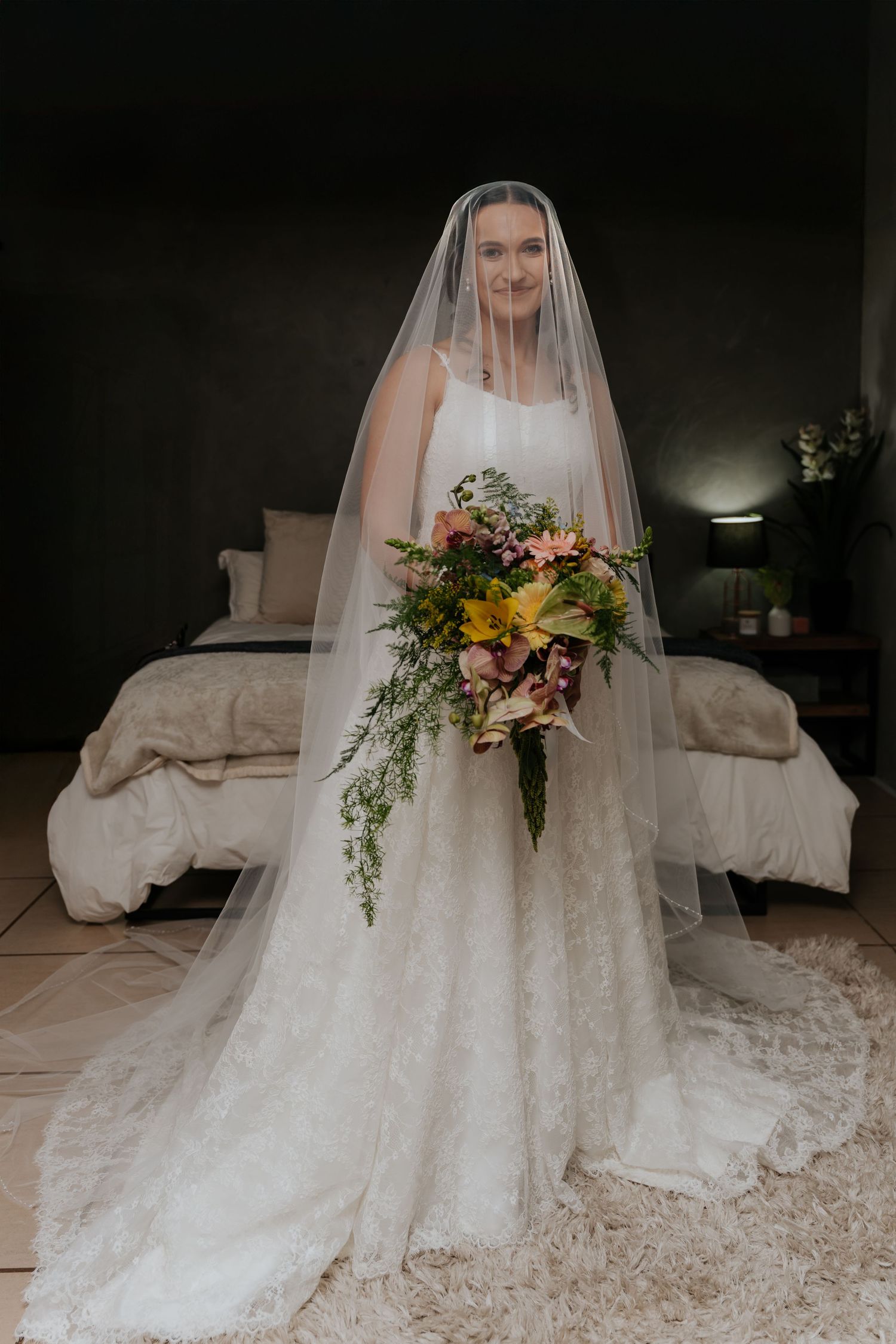 A bride in a flowing lace wedding gown and veil holds a vibrant cascading bouquet with yellow flowers and greenery.