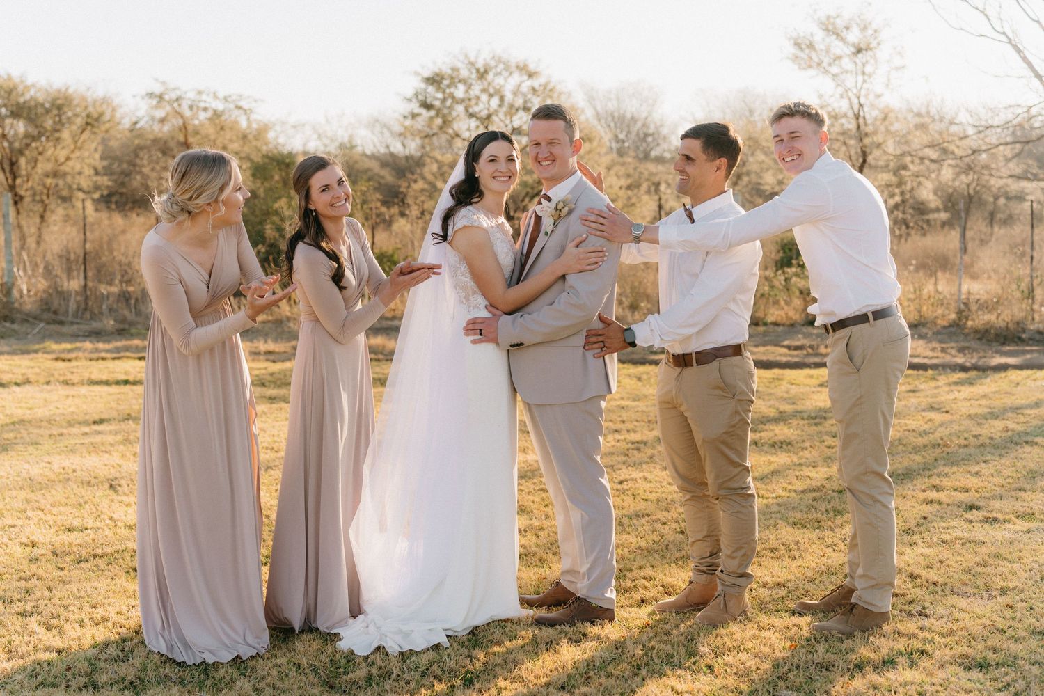 Wedding party in neutral tones poses together during golden hour in a natural outdoor setting.