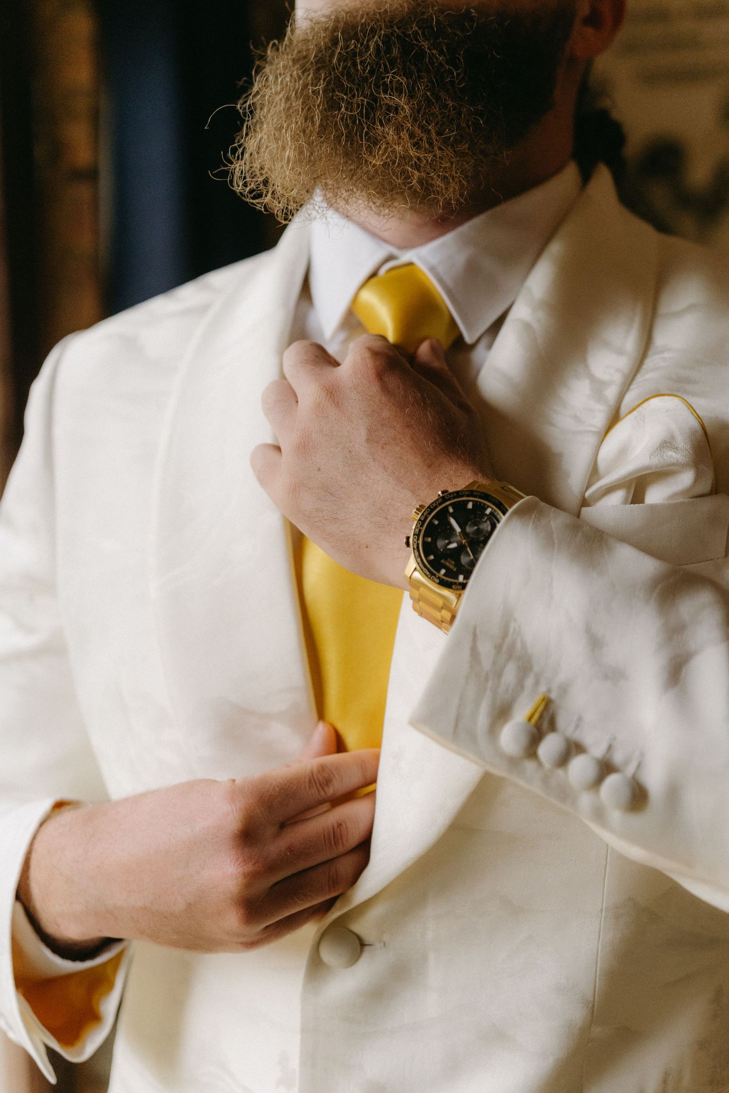 Close up of person adjusting yellow tie with white suit and gold watch, formal wedding attire details.
