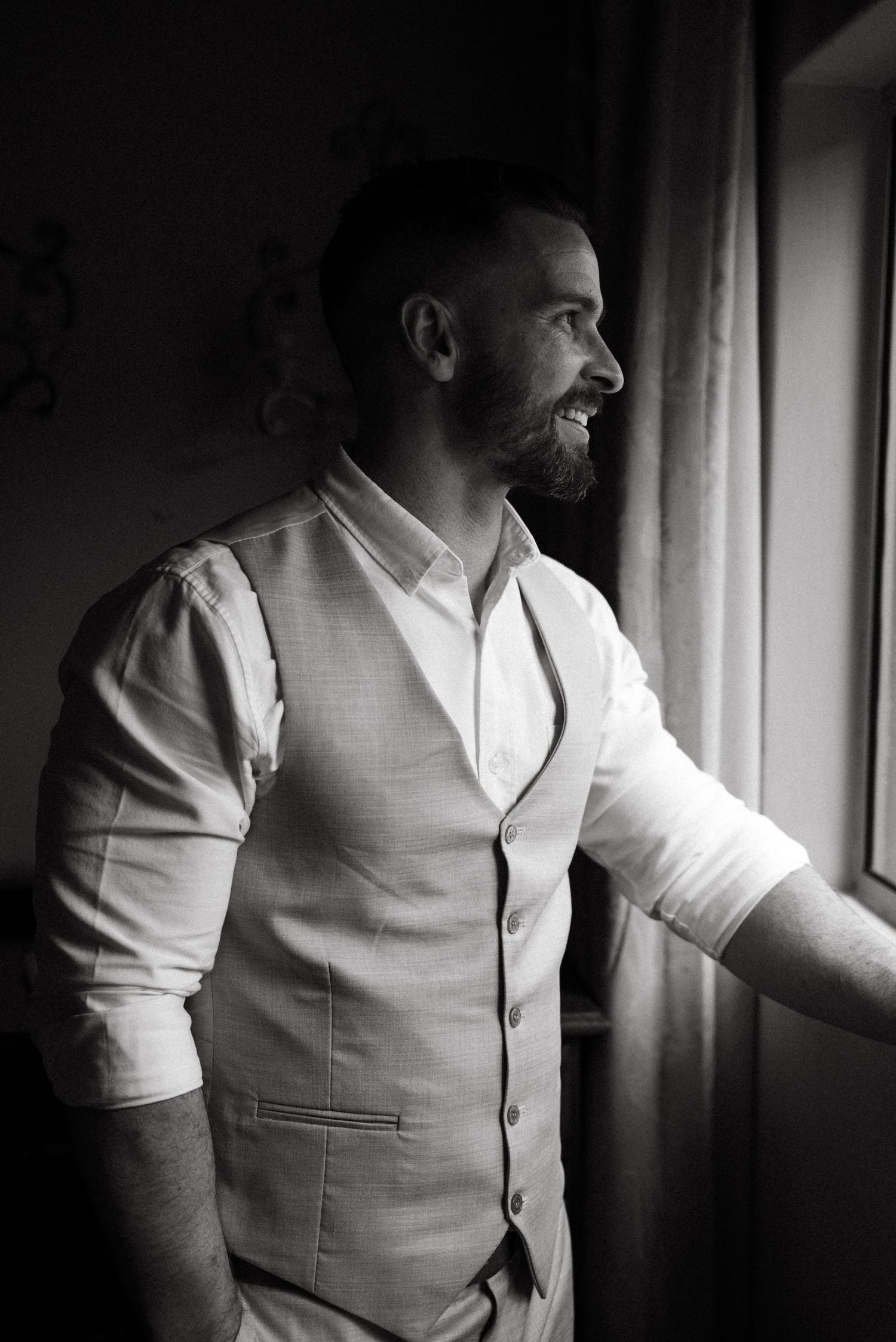 A black and white portrait of a bearded man in a white dress shirt and vest gazing thoughtfully out a window.