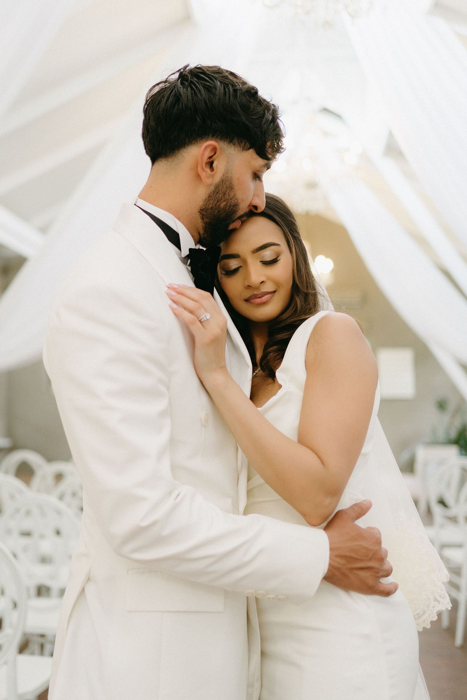 A tender moment between newlyweds in white attire against a light background.