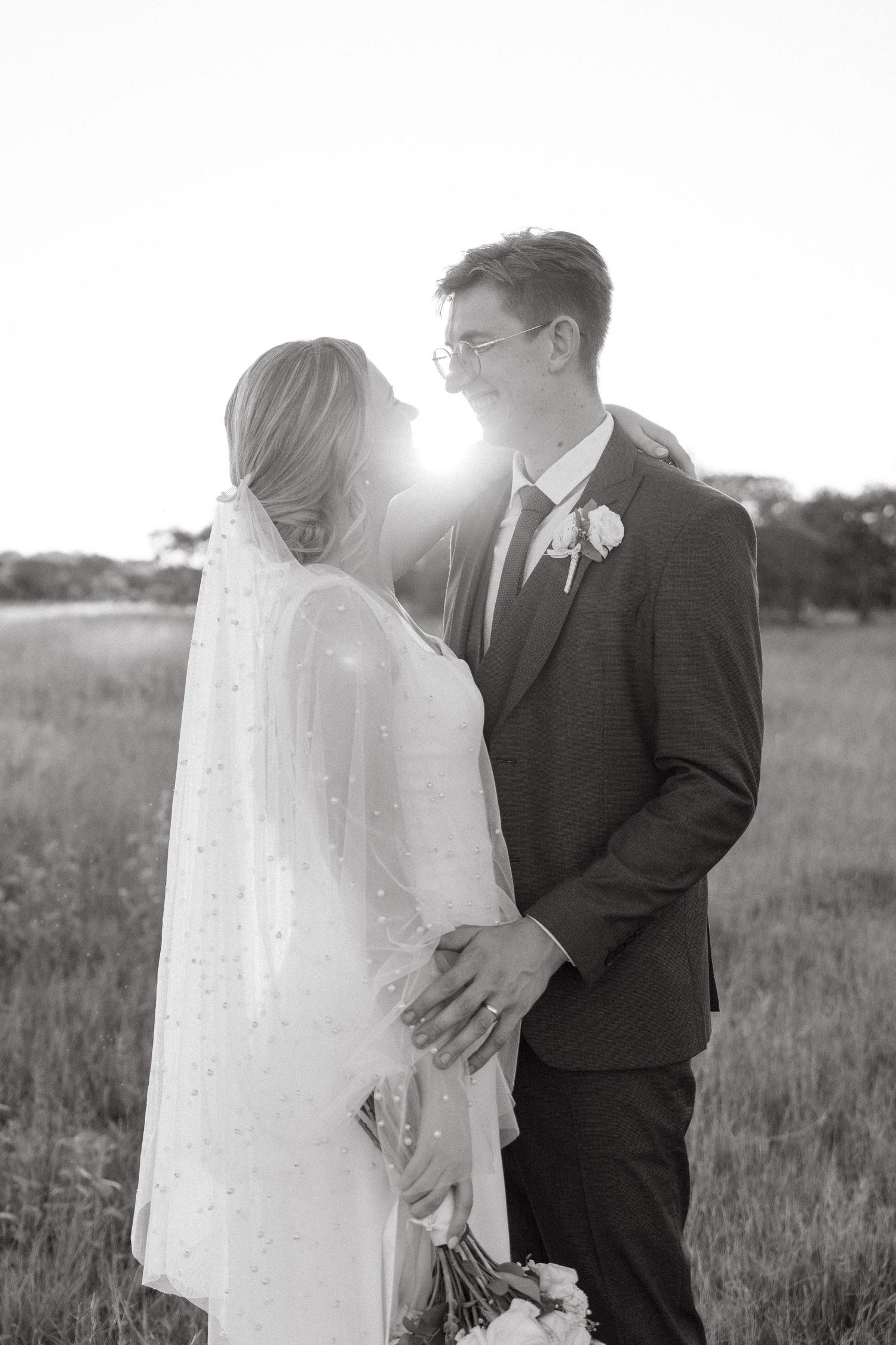A romantic black and white wedding photo of newlyweds embracing in a sunlit field with long grass.
