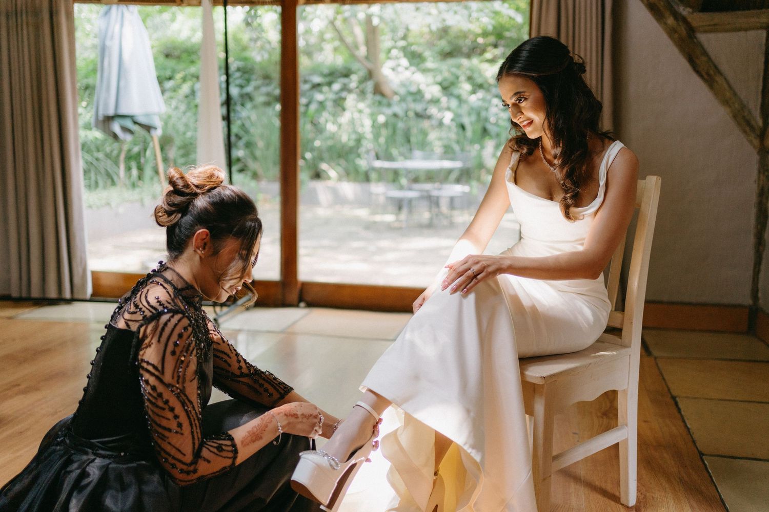 A bride in white dress sits in a chair while someone helps with her shoes during wedding preparations.