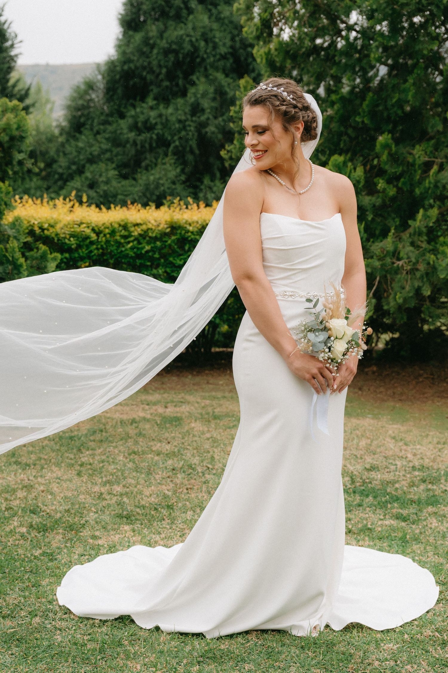 Elegant bride in fitted white gown with flowing veil poses in garden setting.