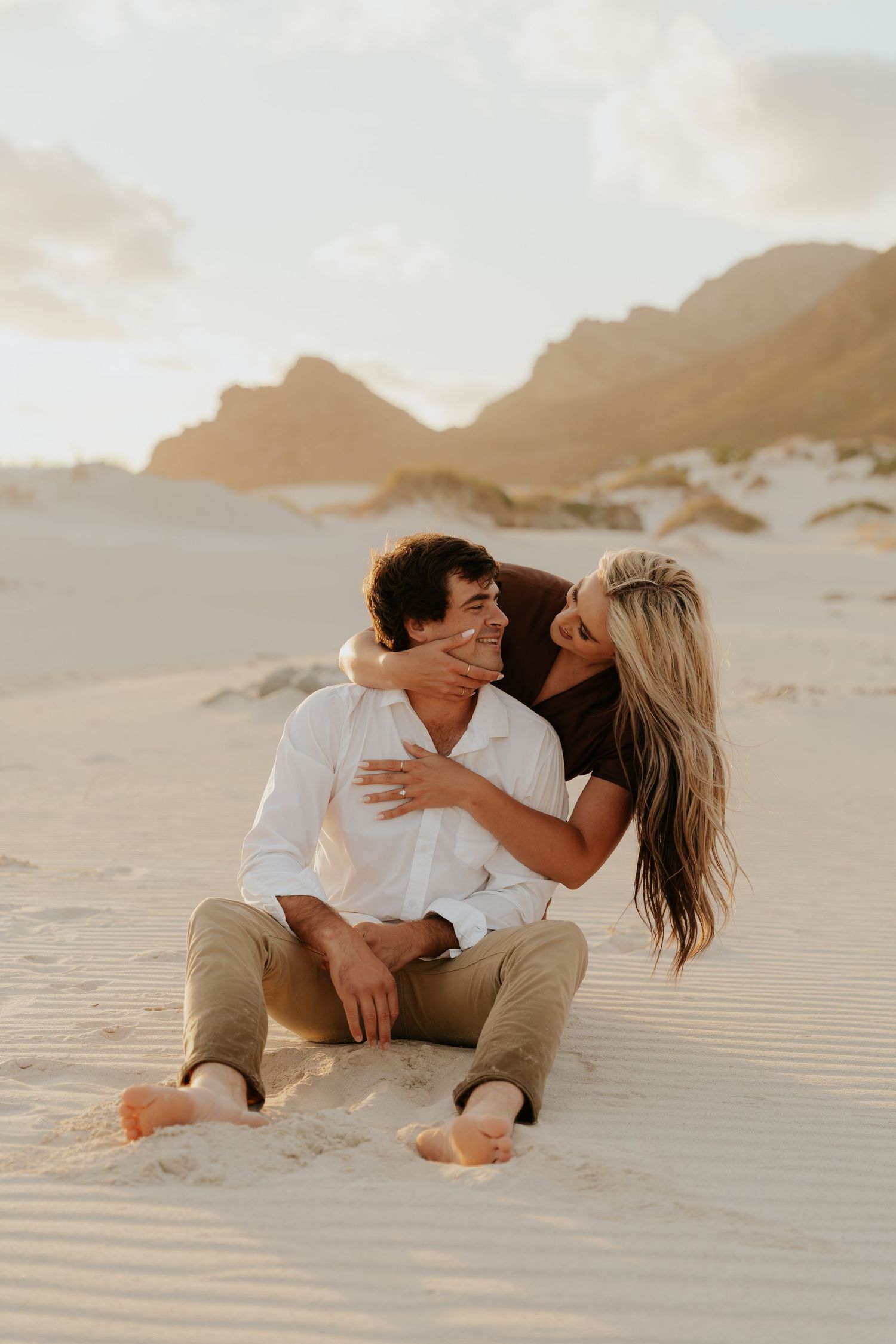 Couple sitting on sandy beach dunes, woman embracing man from behind, smiling, with mountains in golden hour light.