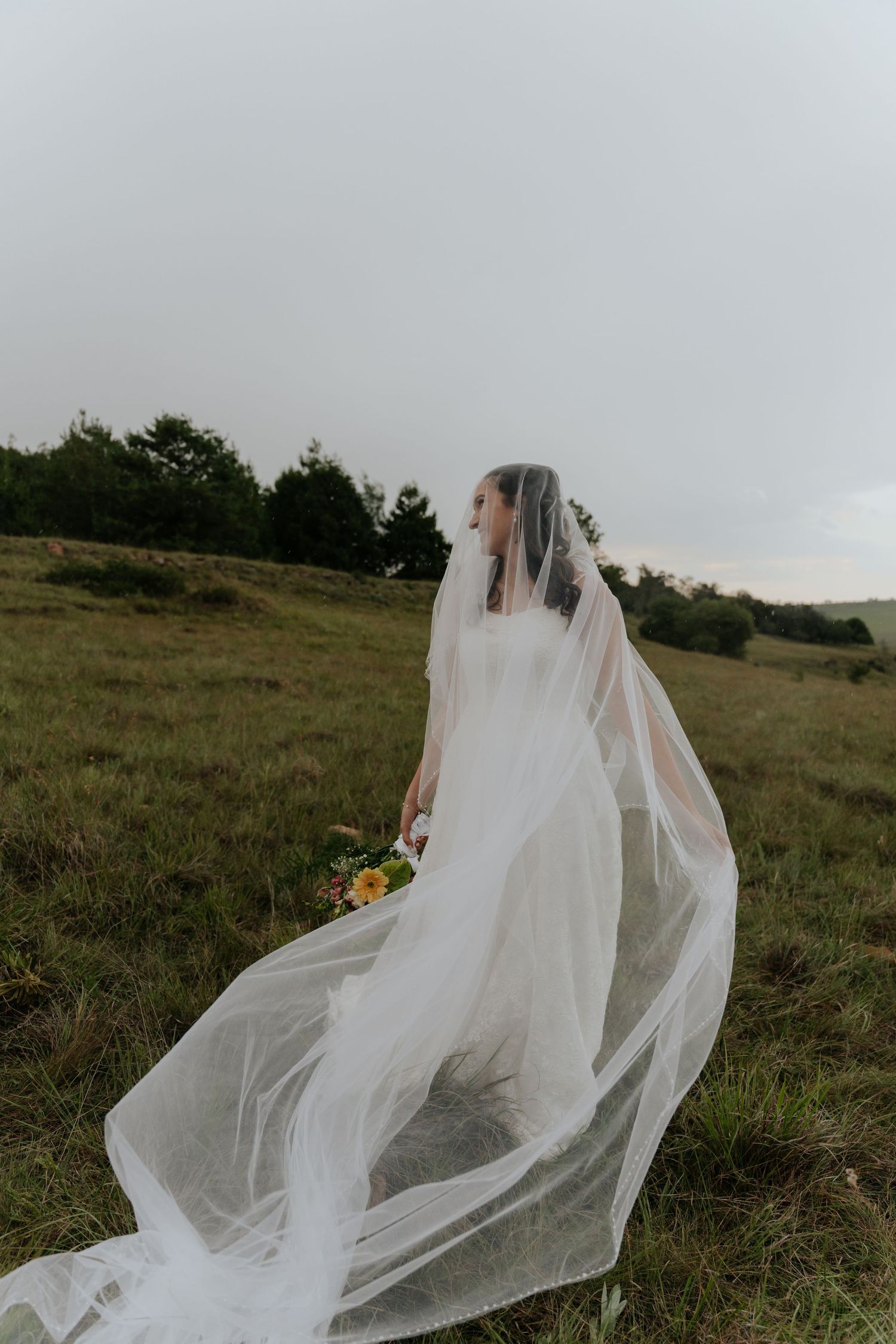 A bride in a flowing white dress and long veil stands in a grassy meadow holding a small bouquet of flowers.