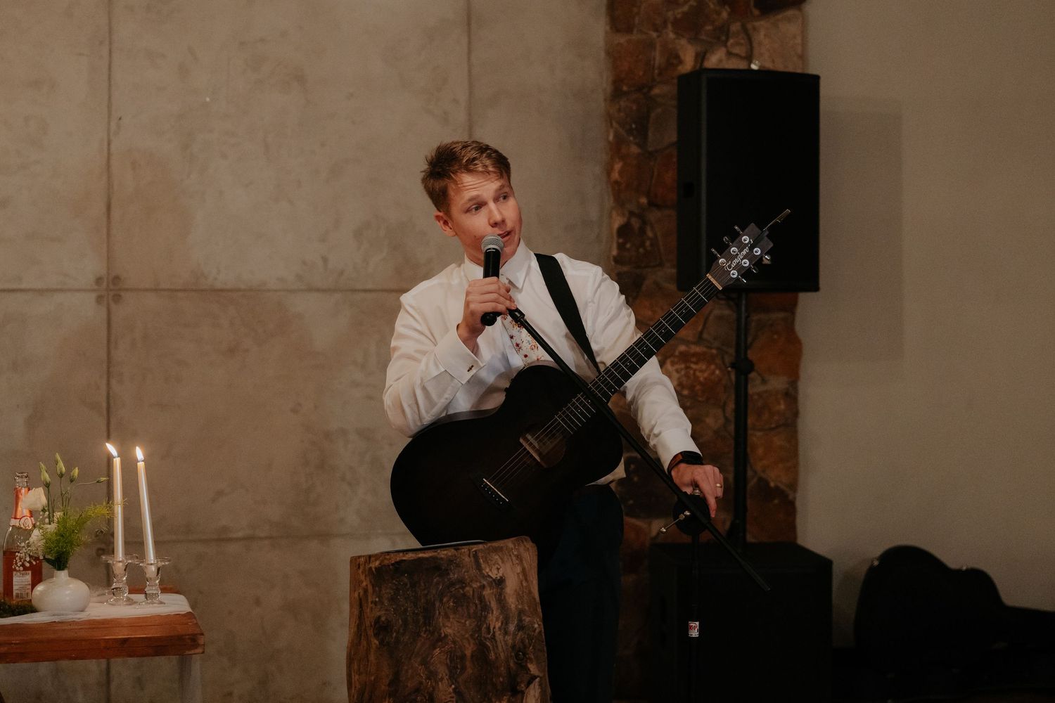 Musician in white shirt plays acoustic guitar while seated on rustic wooden stump in dimly lit venue.