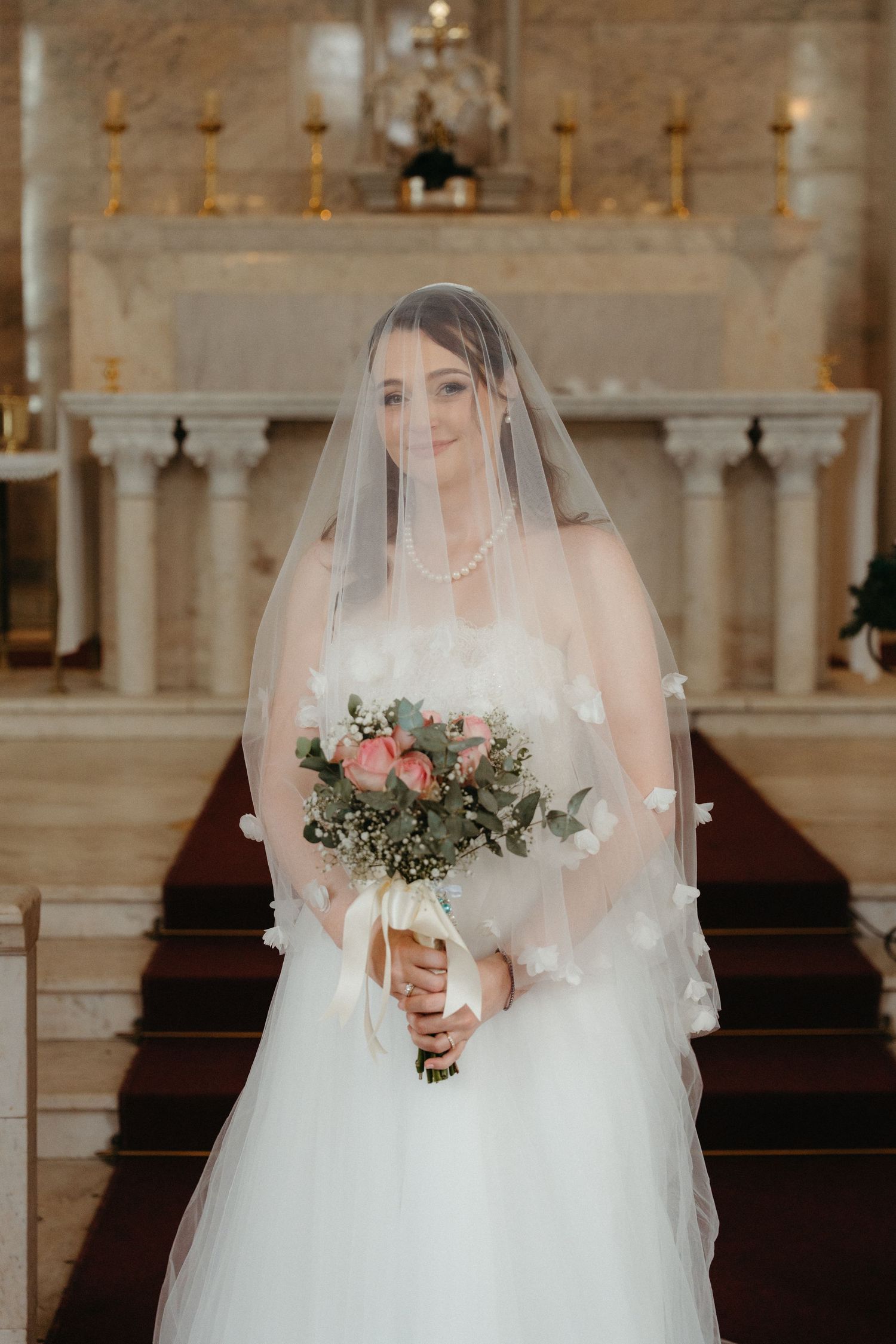 A bride in a white wedding dress and veil holds a bouquet while standing in Nazareth House Chapel
