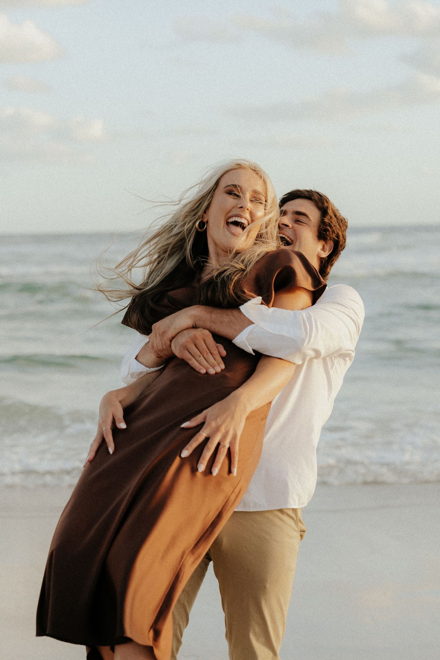 A joyful couple embracing and laughing together on a sandy beach with ocean waves in the background.