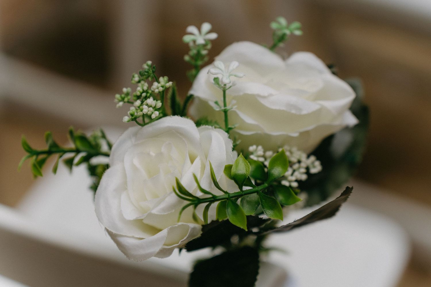 A delicate white rose boutonniere with small greenery and silver wire wrapped stem rests against a blurred background.