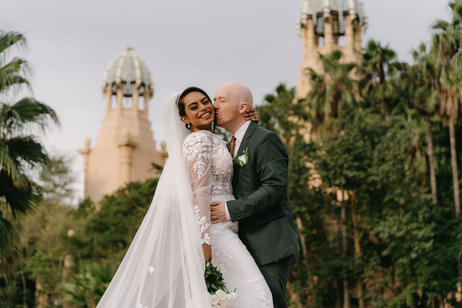 A couple in formal wedding attire share romantic moments in a palm tree-lined garden near cathedral architecture.