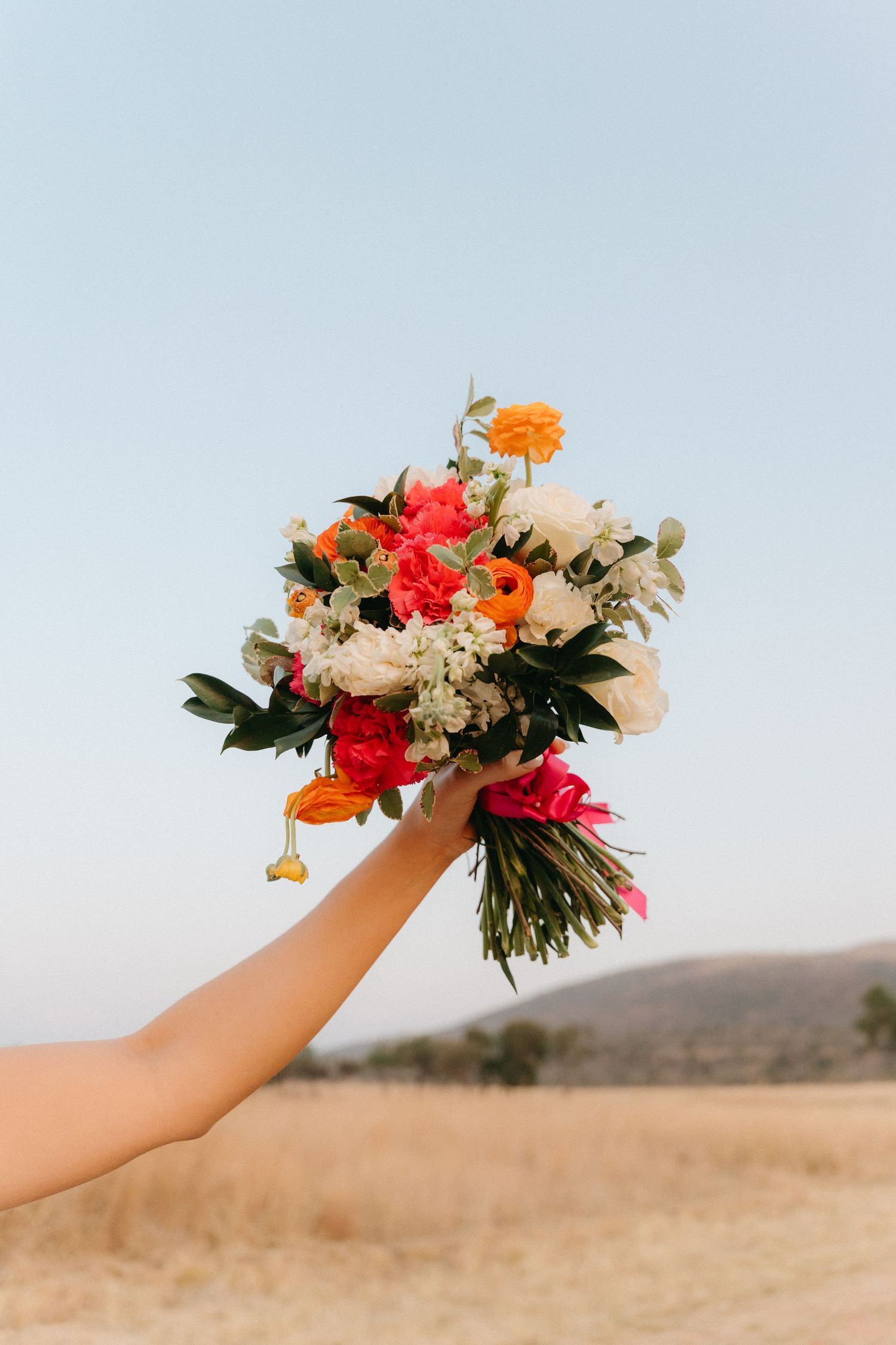 Colorful bridal bouquet with orange and red flowers held against a desert landscape backdrop.