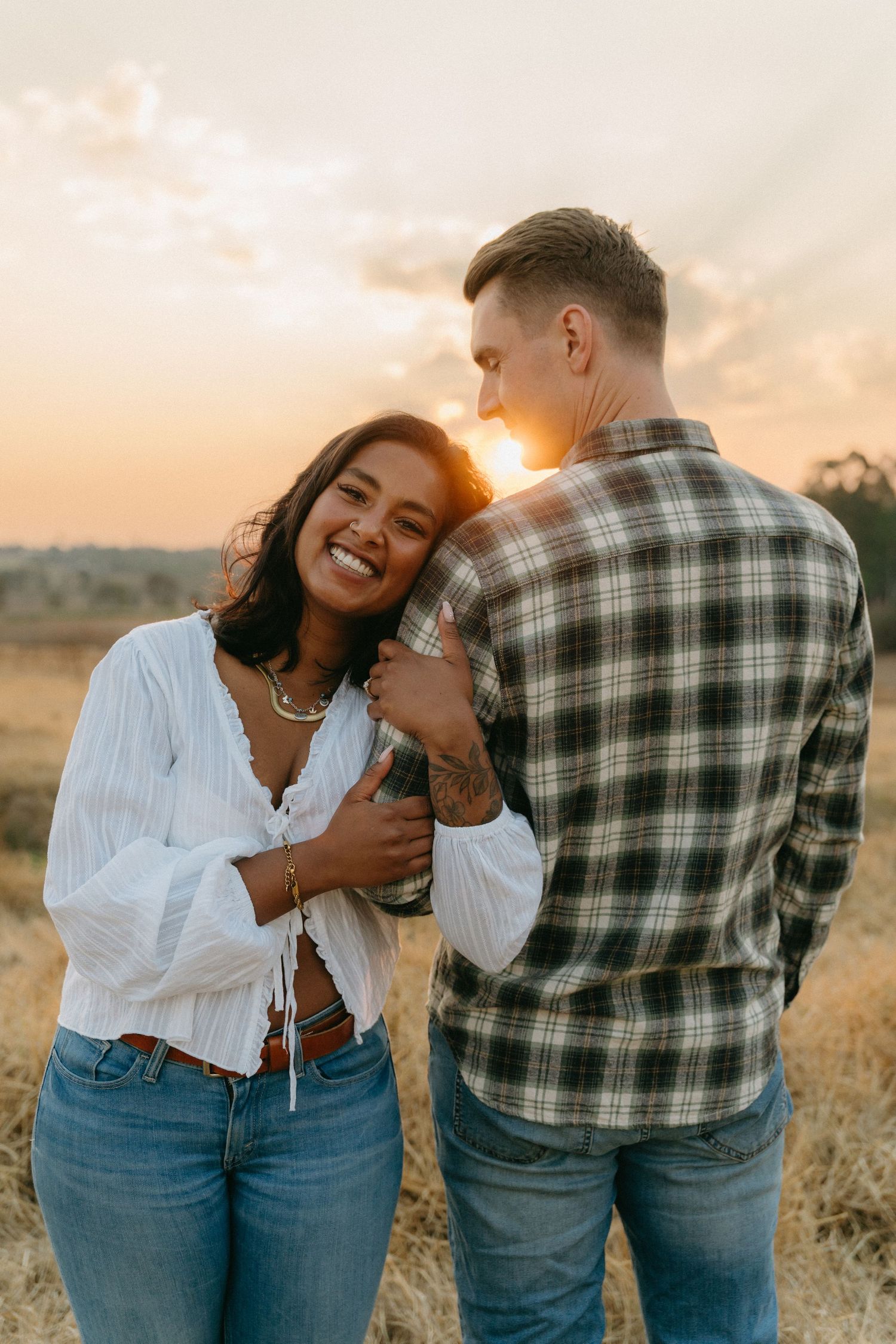 Couple embracing in golden sunlit field wearing matching denim and plaid during sunset photoshoot.