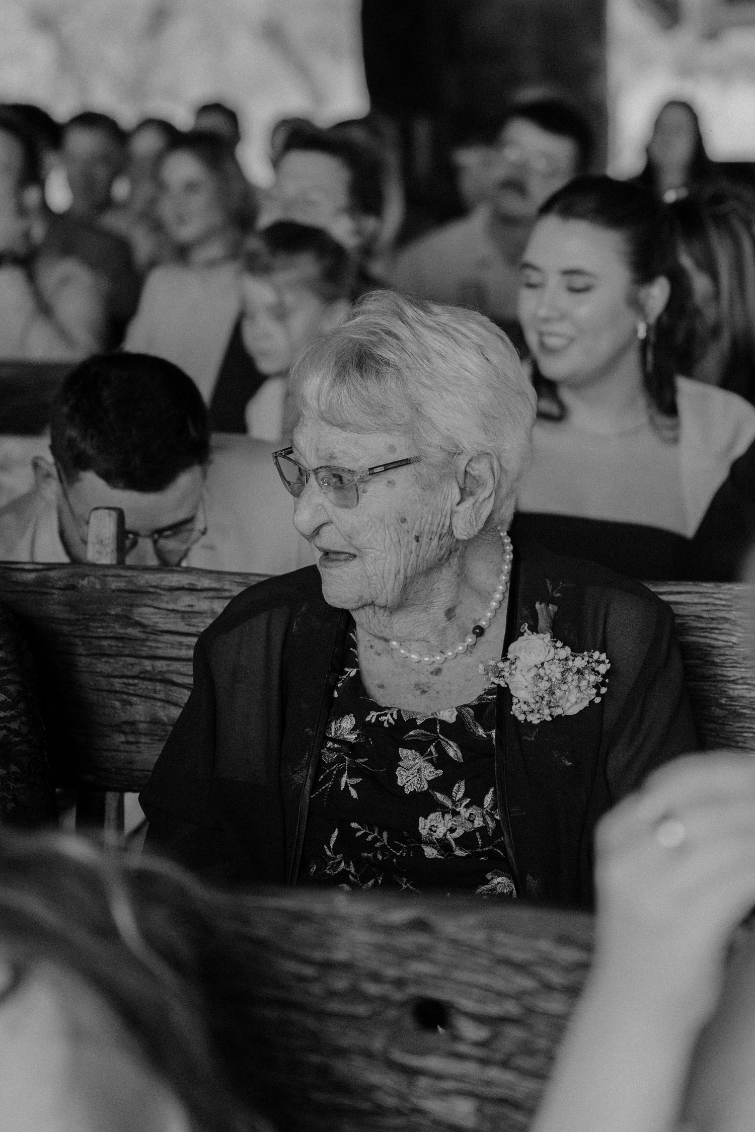 An elderly guest in a floral dress and glasses watches the wedding ceremony from a wooden pew.