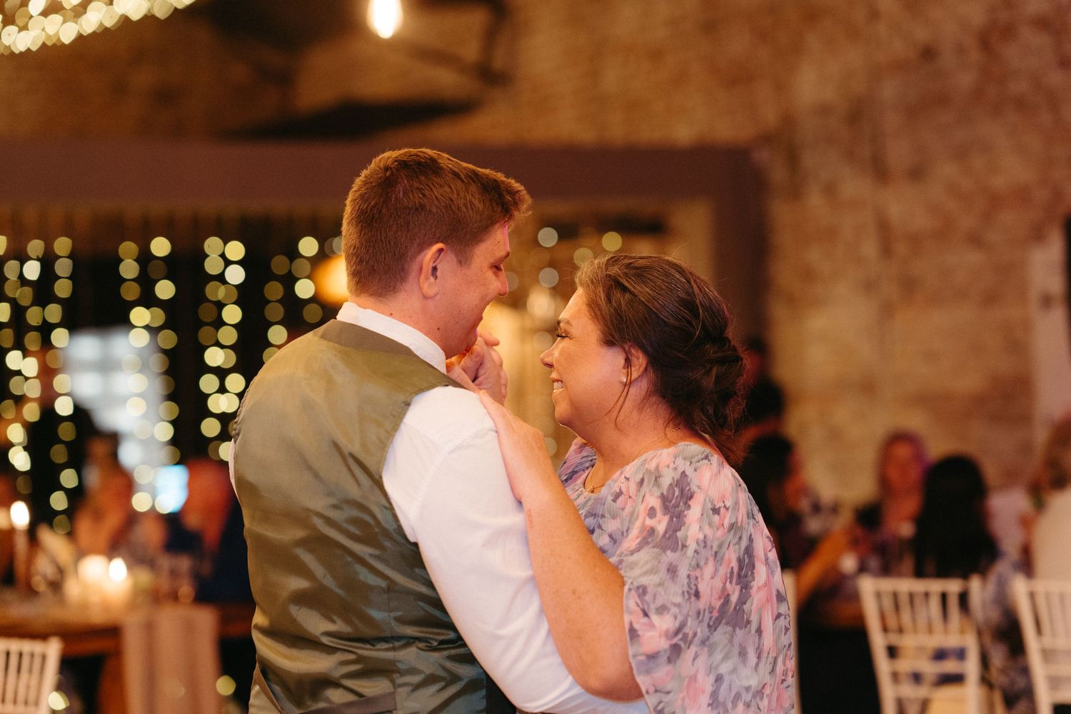 A couple shares a romantic dance moment together under twinkling string lights at an indoor wedding reception venue.