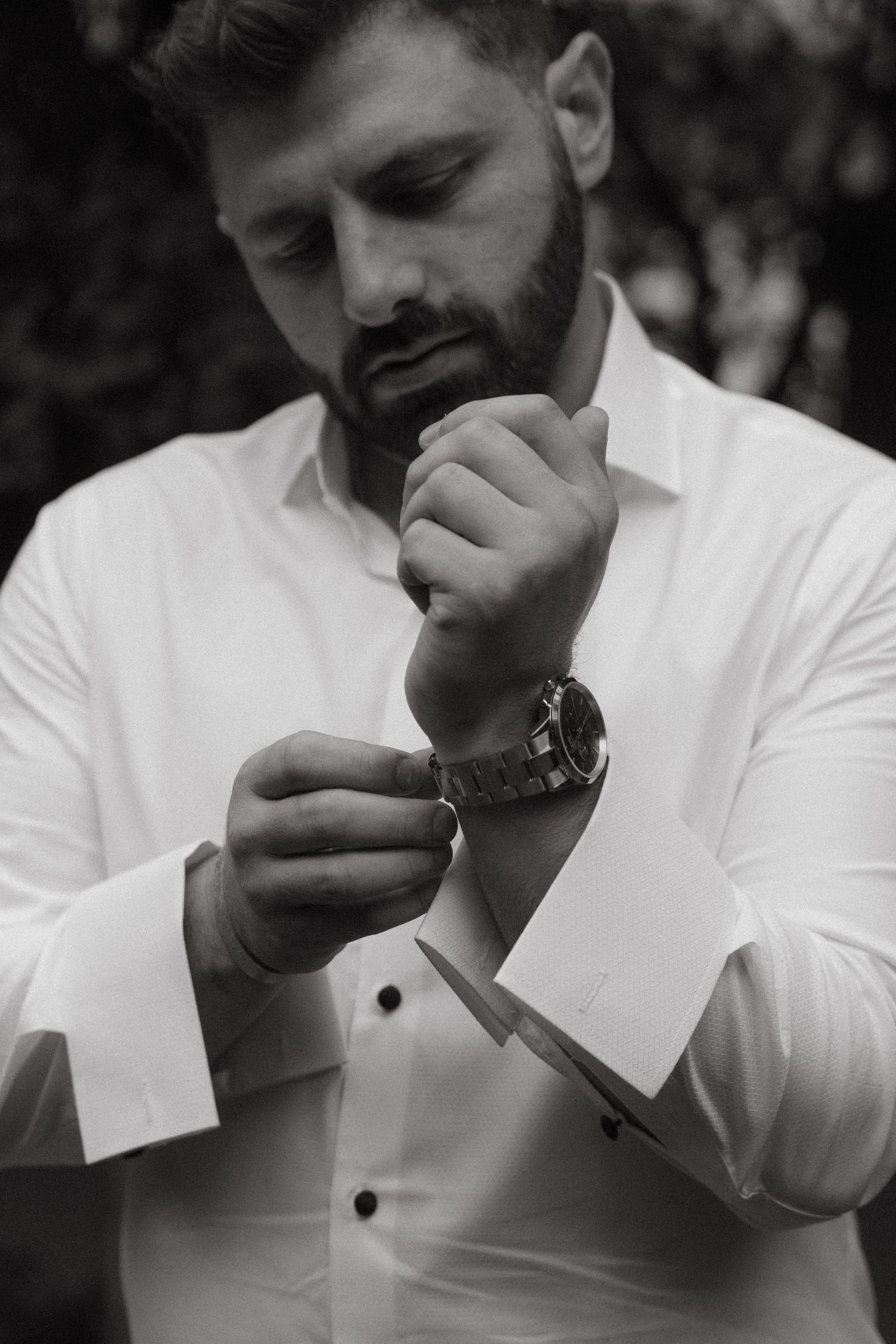 Black and white portrait of bearded groom in white dress shirt fastening cufflinks outdoors in Johannesburg