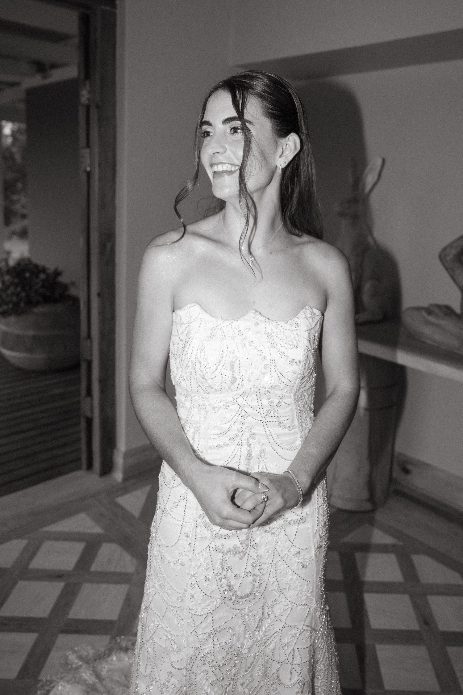 Black and white photo of a smiling bride in a strapless beaded wedding gown, hands clasped, standing in an elegant venue.
