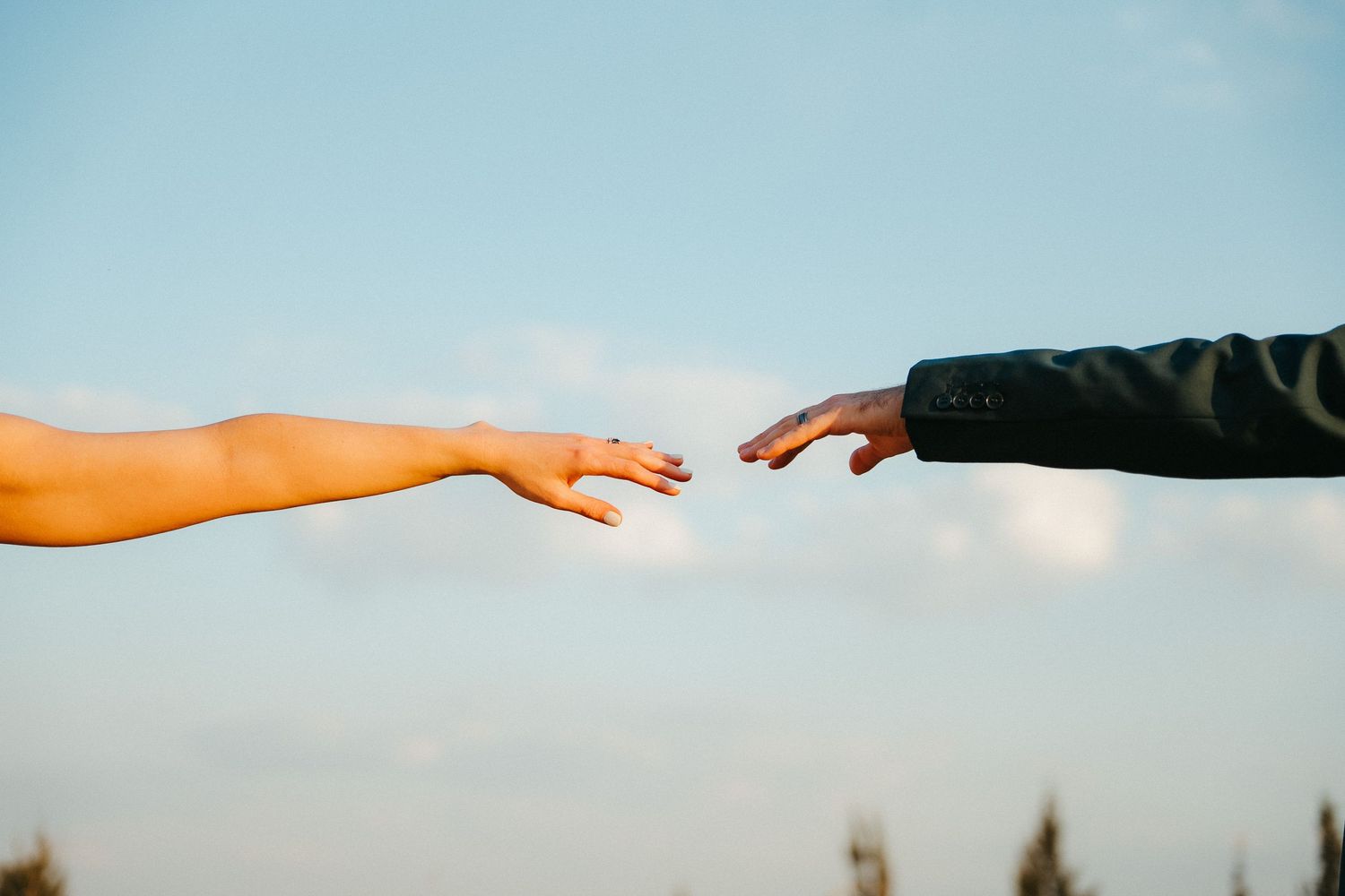 Two hands reaching toward each other against a blue sky, symbolizing connection and unity.