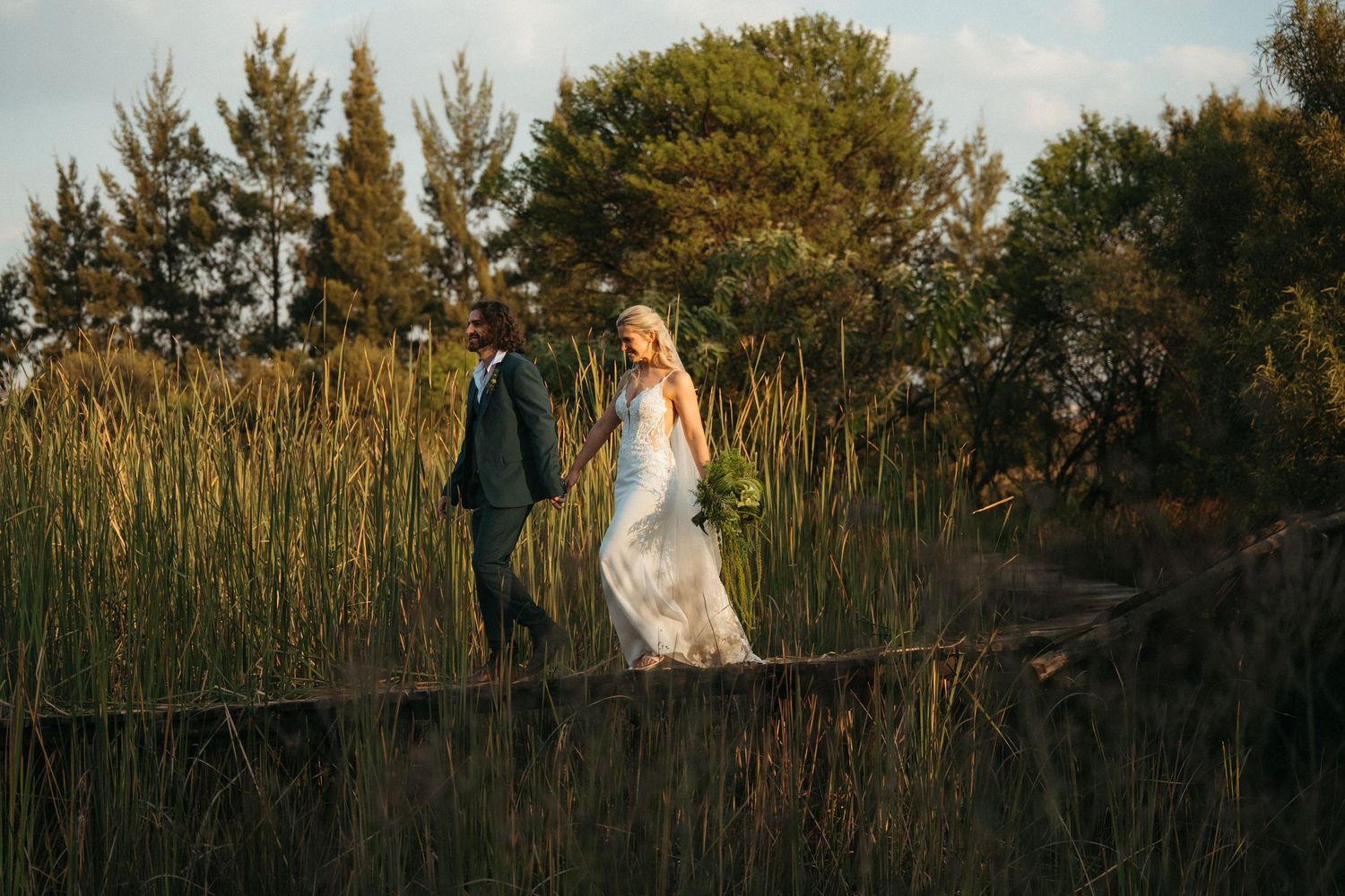 Bride and groom walk hand in hand on a wooden path surrounded by tall golden grass and lush green trees.
