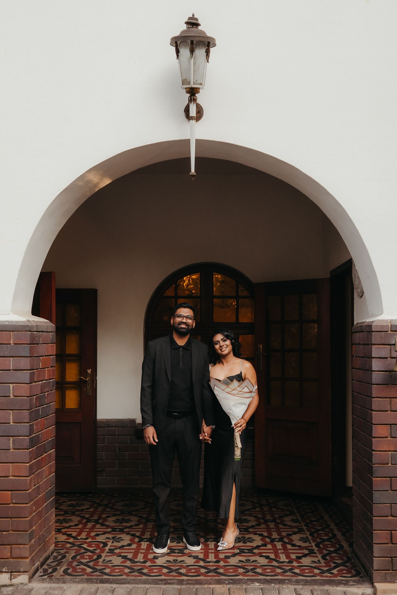 Couple posing under an arched doorway, man in black suit, woman in silver dress, brick entrance with vintage lantern.