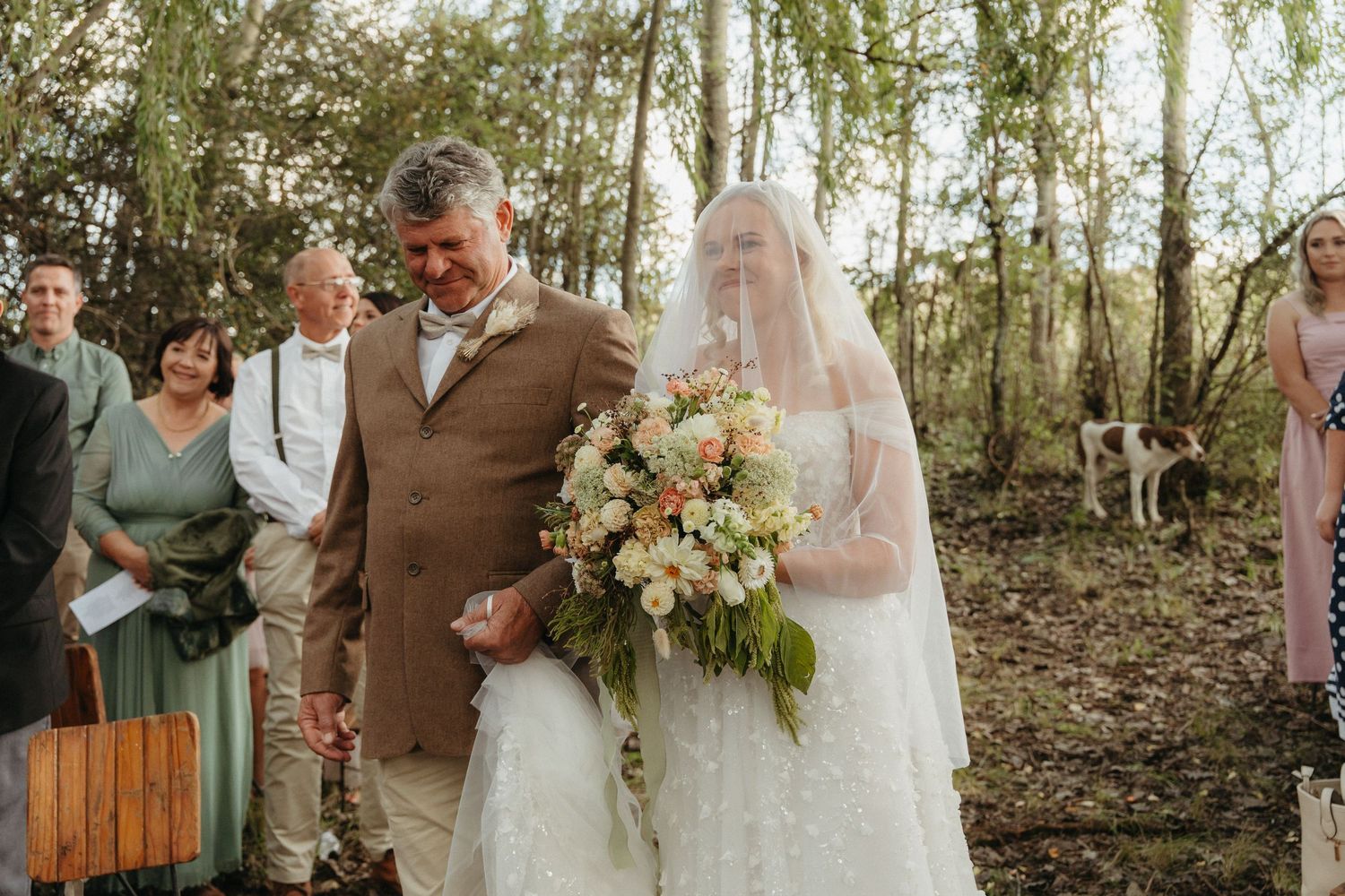 Bride in white lace gown and veil walks with man in brown suit through woodland wedding aisle holding peach floral bouquet.