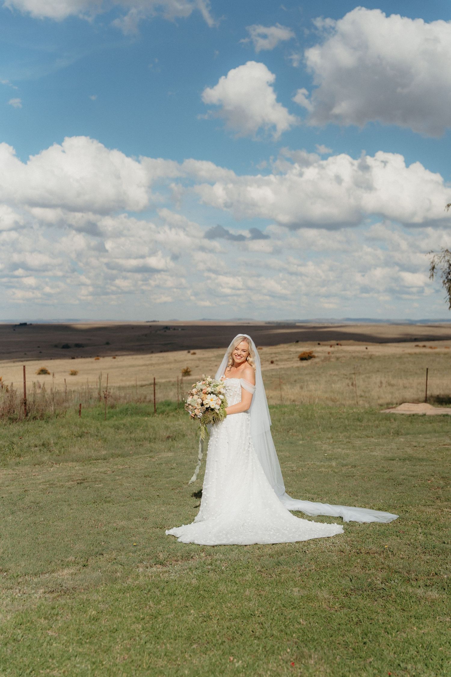 Bride in white gown and veil holding bouquet stands in green field under blue sky with rolling hills backdrop.