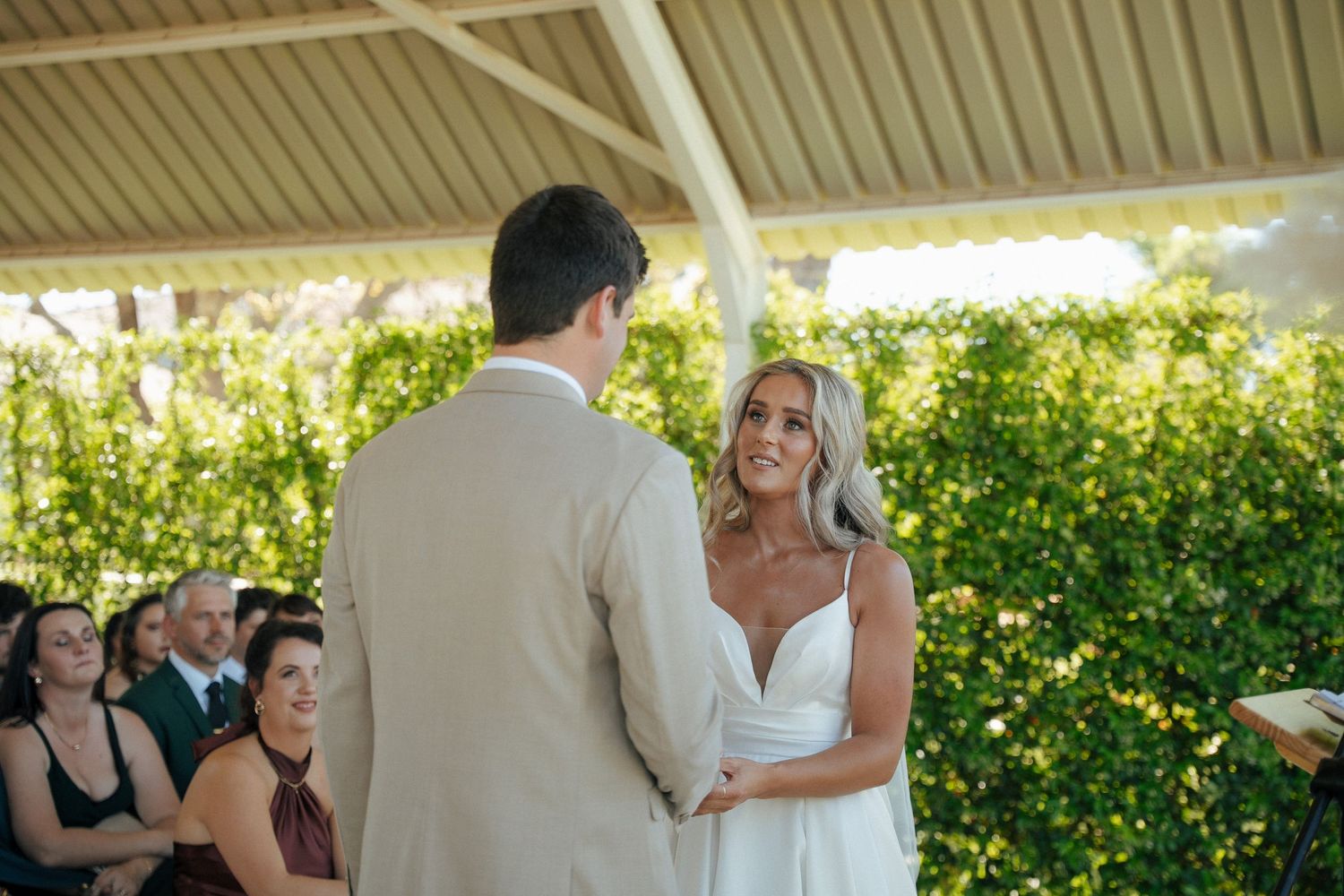 Bride in a white dress holds hands with the groom in a beige suit during an outdoor wedding ceremony as guests watch under a pavilion.