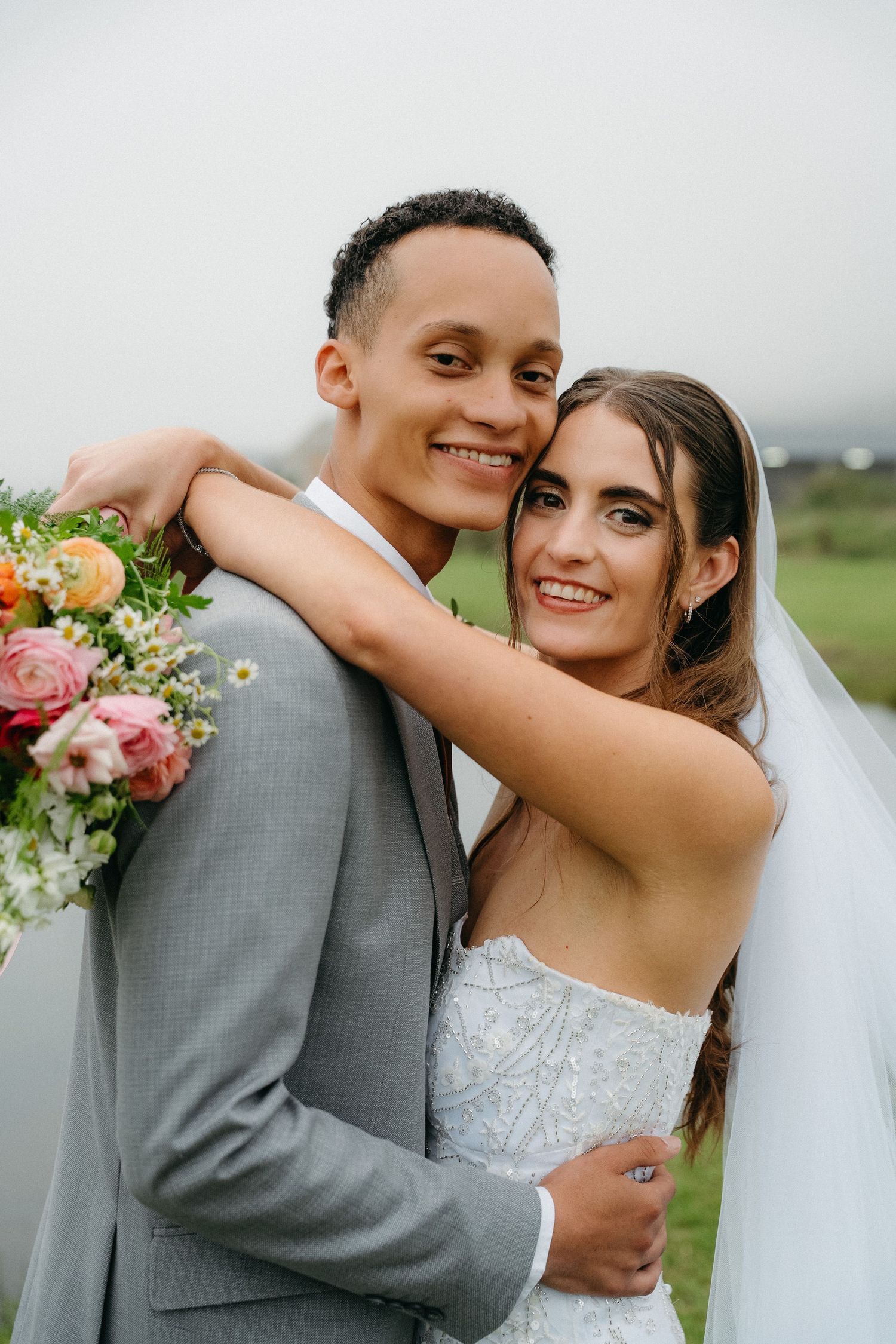 A smiling bride and groom pose together outdoors, she wearing a white gown with veil, he in a gray suit, holding colorful flowers.